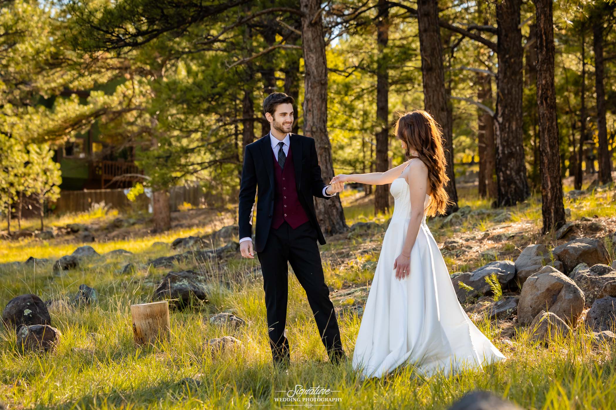 Photo by Tavits Photography A couple in formal attire, the man in a dark suit and the woman in a white wedding dress, hold hands and stand together in a sunlit forest clearing surrounded by trees and rocks.