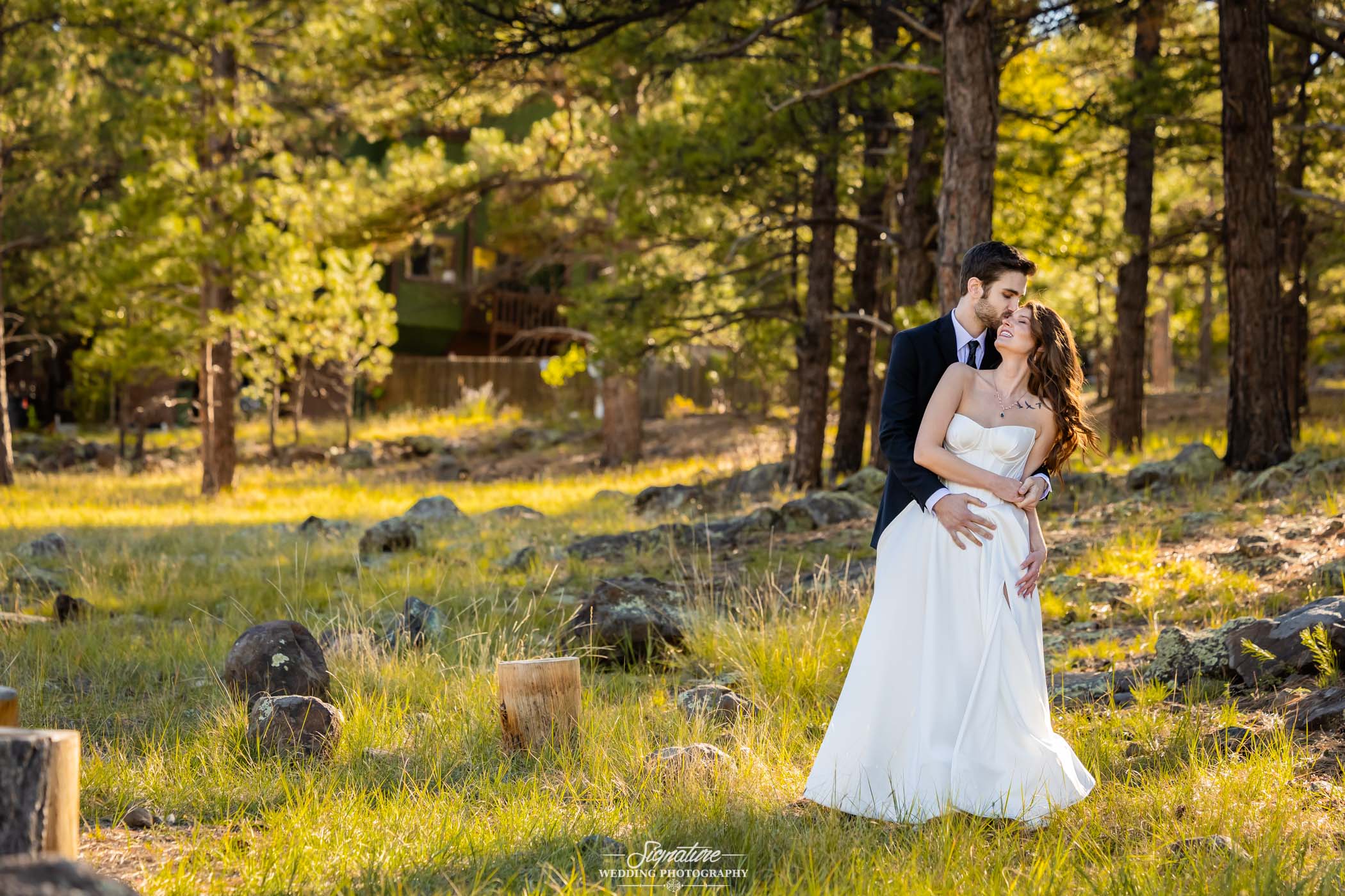 Photo by Tavits Photography A bride and groom embrace and smile at each other in a sunlit forest clearing, surrounded by tall trees and green grass. The bride wears a white dress and the groom is in a dark suit.