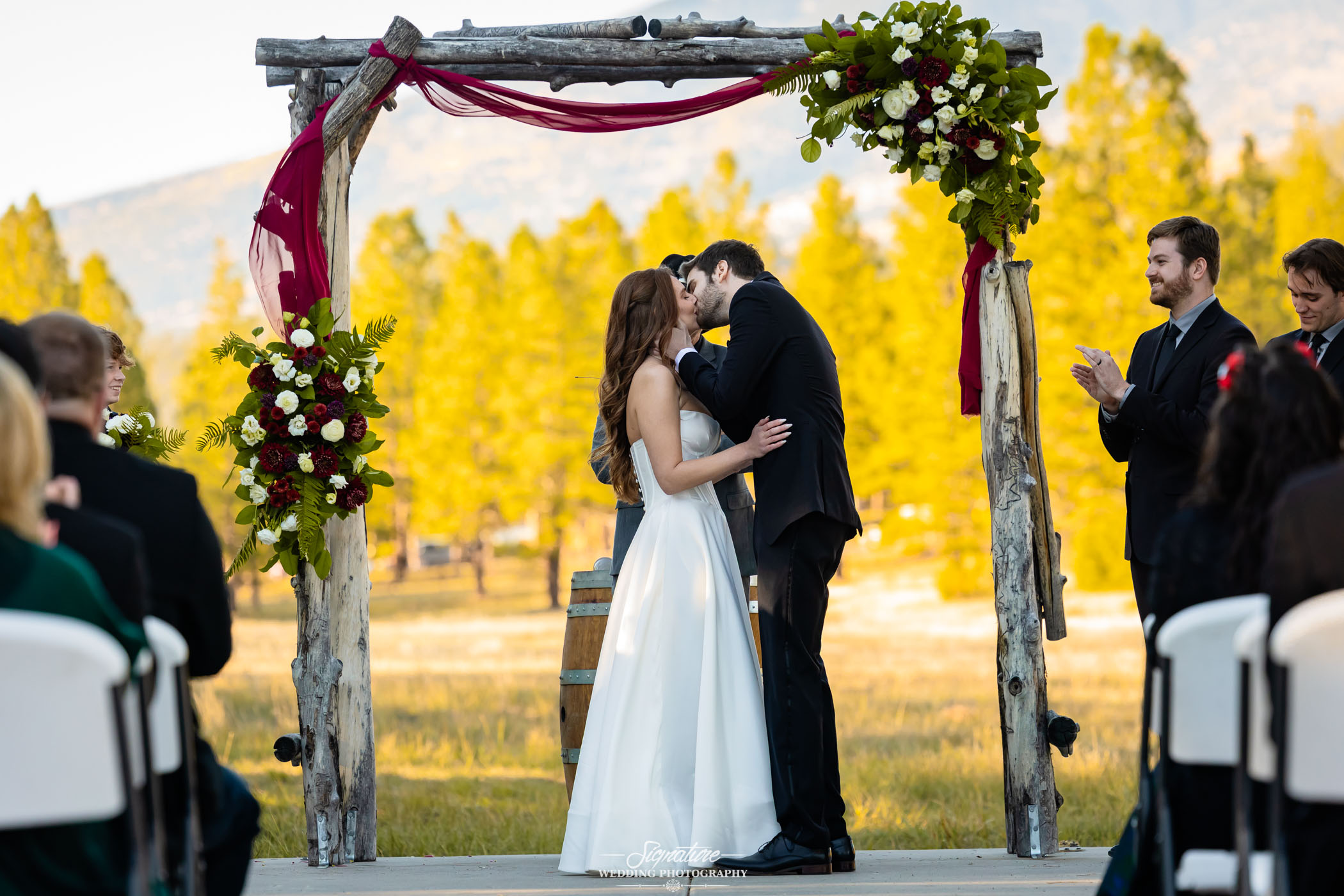 Photo by Tavits Photography A bride and groom share their first kiss under a rustic wooden wedding arch decorated with flowers and burgundy fabric, surrounded by guests seated outdoors in a scenic, sunlit setting.