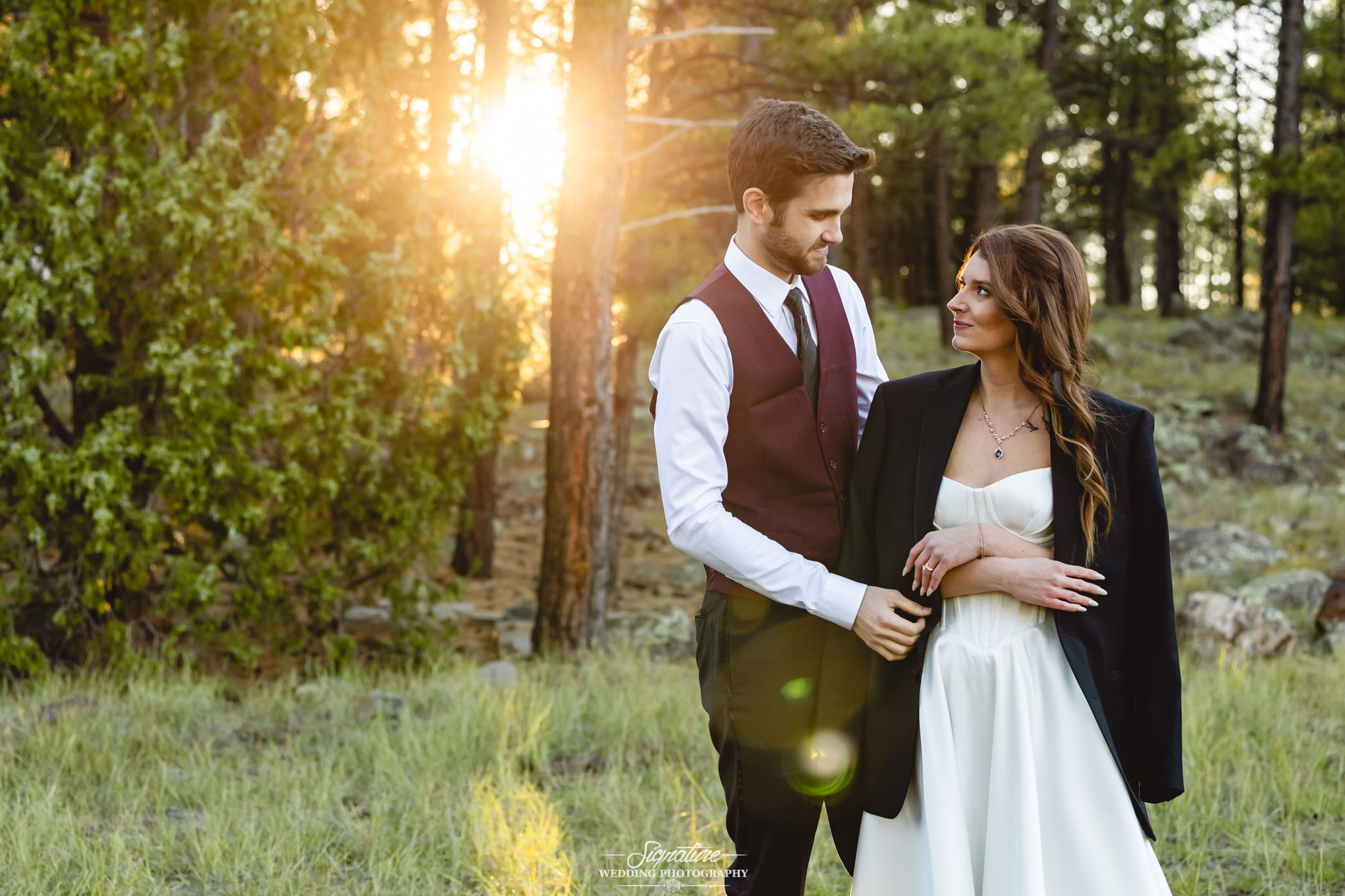 Photo by Tavits Photography A couple stands together in a sunlit forest; the woman in a white dress with a dark jacket draped over her shoulders, the man in formal attire. They gaze at each other, surrounded by trees and green grass as sunlight streams behind them.