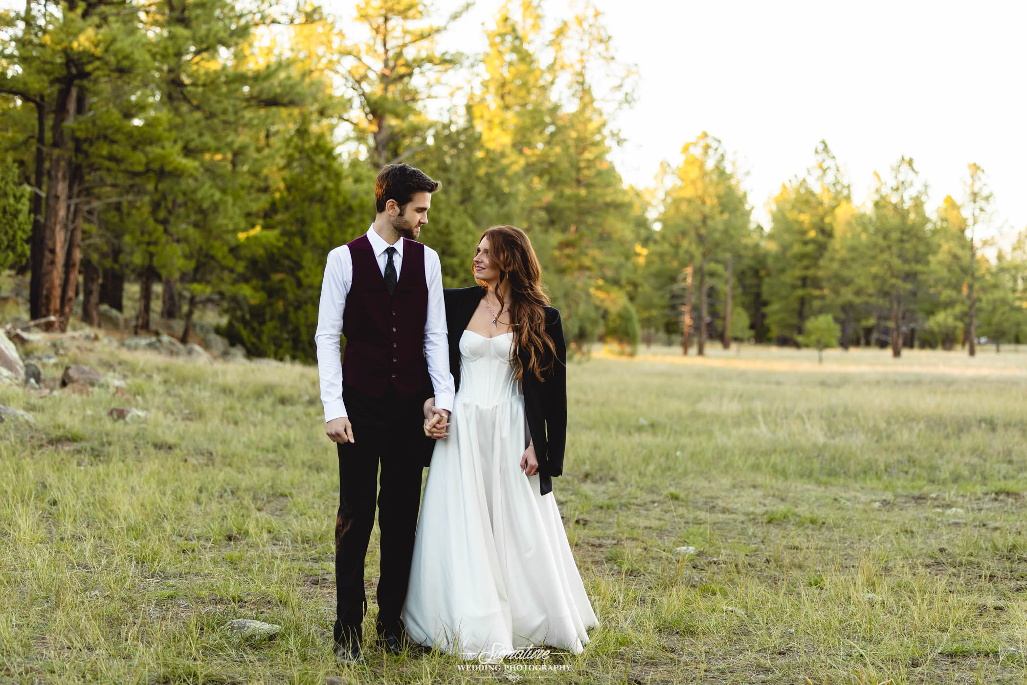 Photo by Tavits Photography A bride and groom hold hands in a grassy field with trees behind them. The groom wears a maroon vest and tie, while the bride wears a white strapless wedding dress and a black jacket.