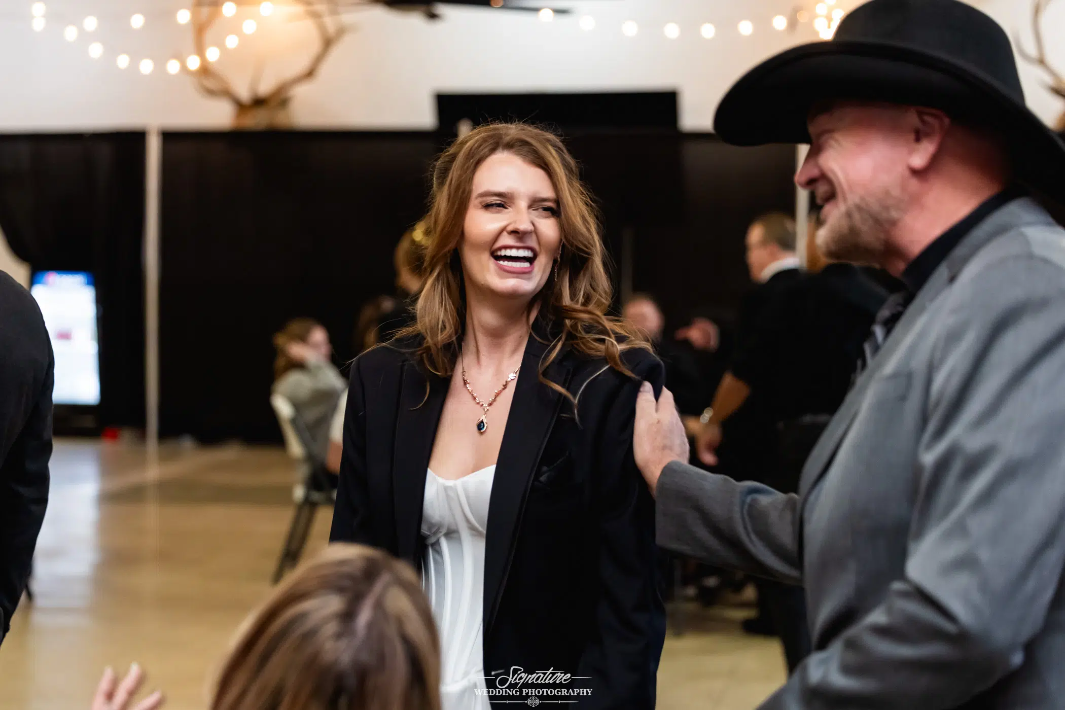 Photo by Tavits Photography A woman in a black blazer and white top smiles and laughs while talking to a man in a cowboy hat at an indoor event with string lights and antler decorations.