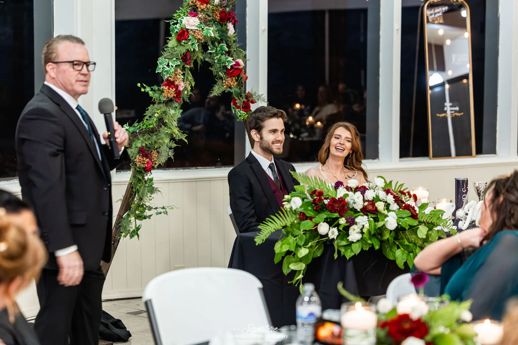 Photo by Tavits Photography A man in a suit speaks into a microphone at a wedding reception, while the smiling bride and groom sit at a decorated table with red and white flowers. Other guests are visible in the background.