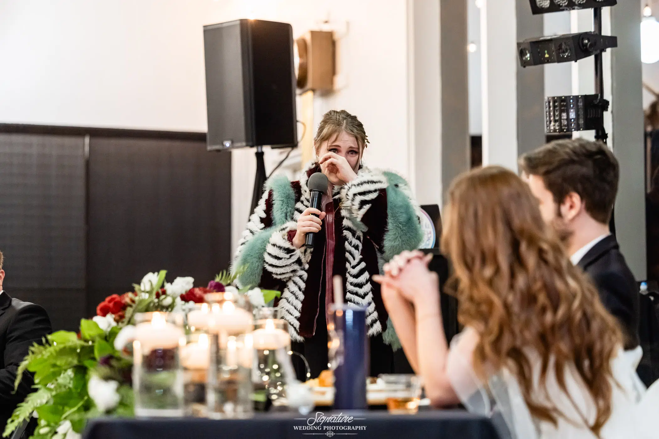 Photo by Tavits Photography A woman in a colorful striped coat stands with a microphone, wiping away a tear while speaking at a wedding reception. Seated guests, floral arrangements, and candles are visible in the foreground.
