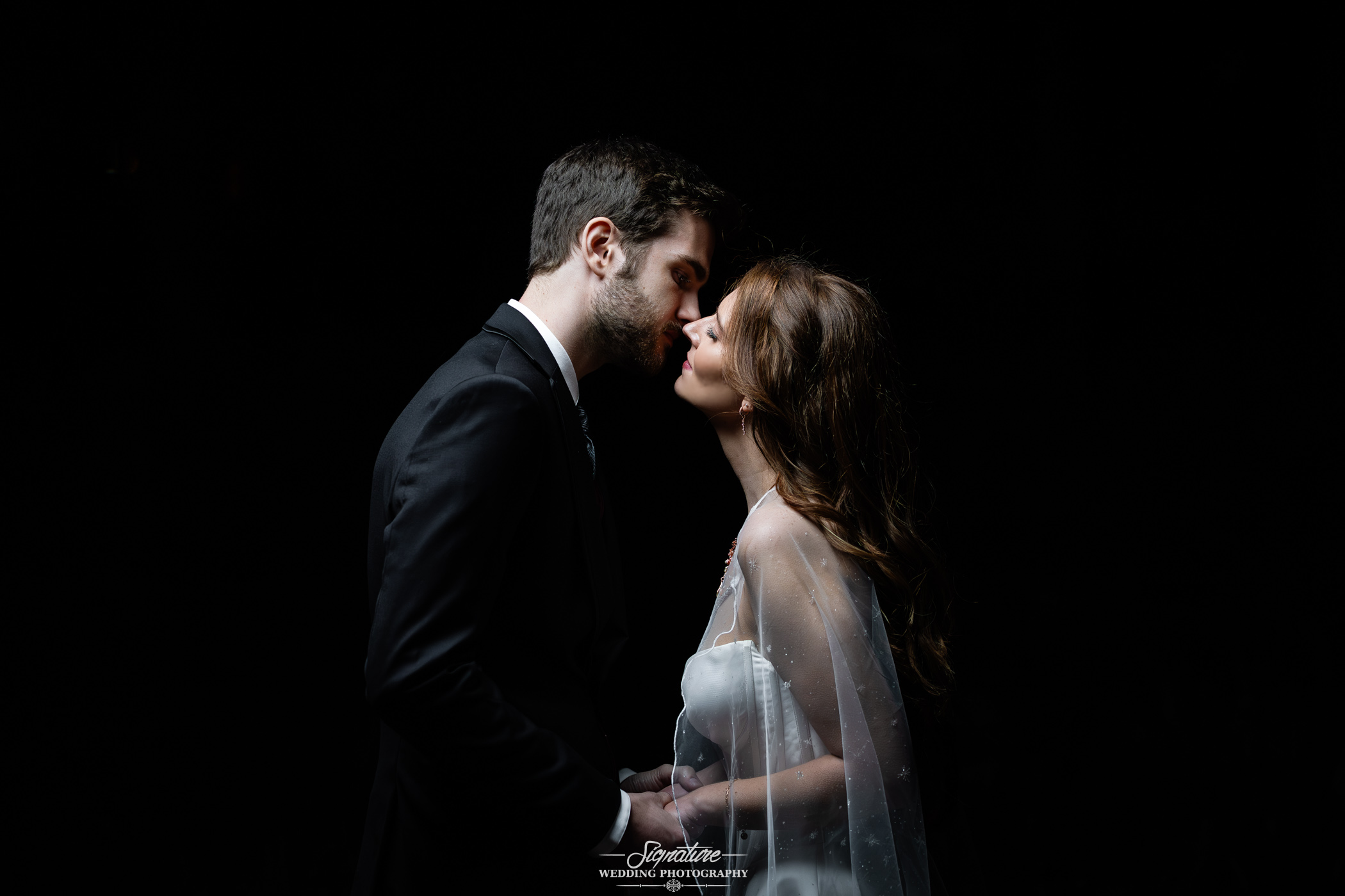 Photo by Tavits Photography A bride and groom stand closely in an intimate pose, holding hands and almost kissing, against a black background. The bride wears a white dress and veil; the groom is in a black suit.