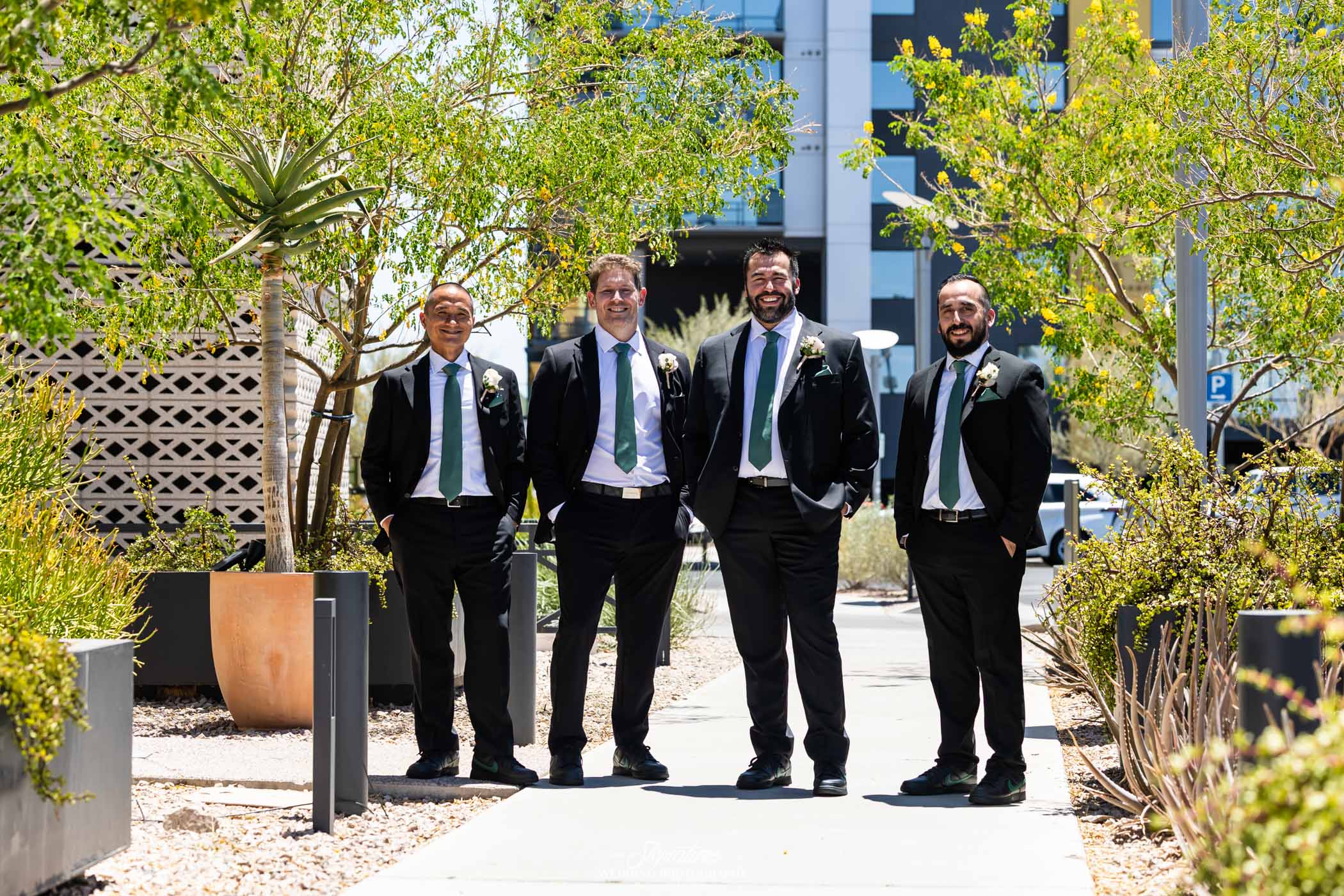 Image by Signature Wedding Photography Four men in suits and ties stand smiling together on a sunny sidewalk surrounded by greenery and modern buildings, suggesting a formal or celebratory occasion.
