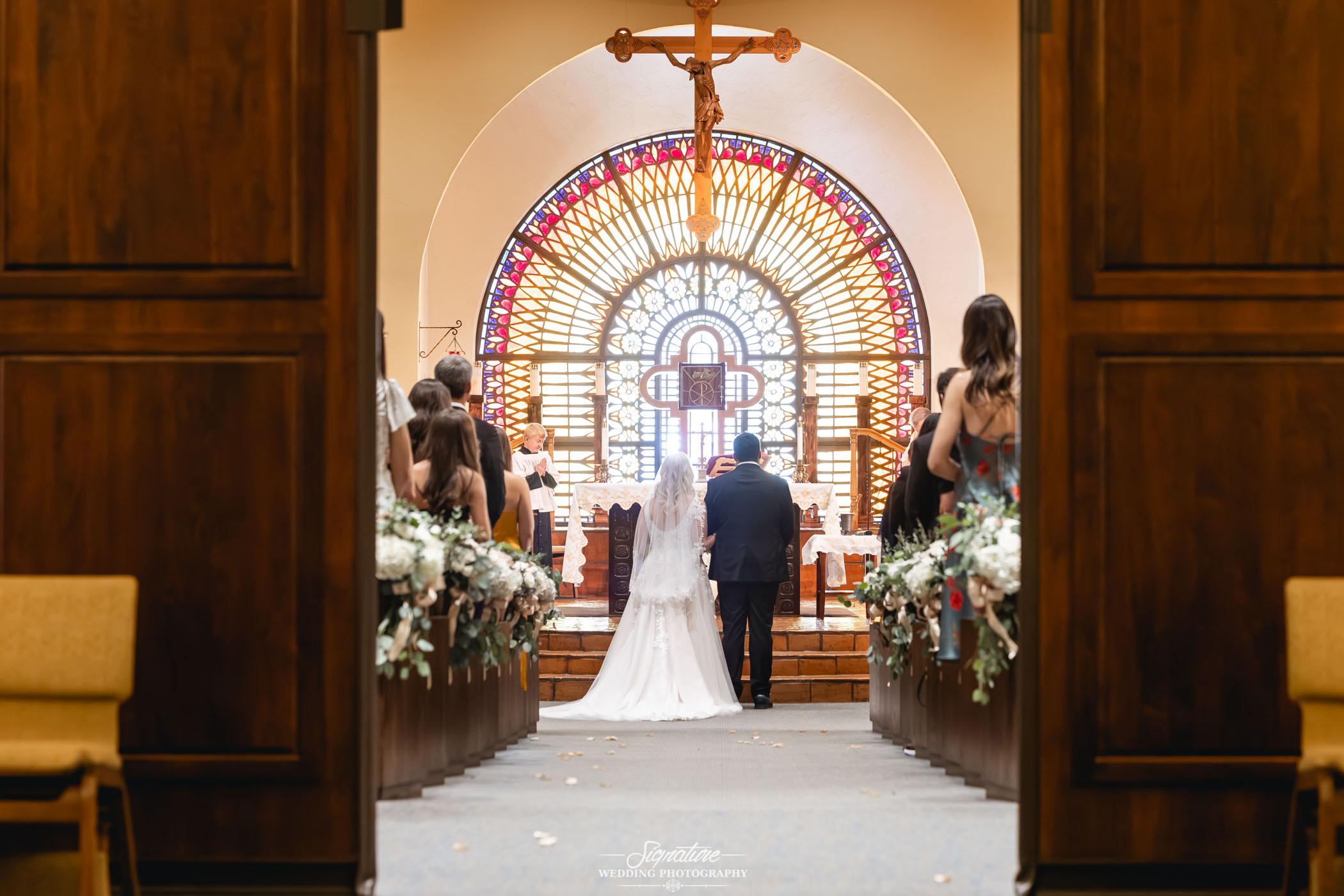 Image by Signature Wedding Photography A bride and groom stand at the altar in a church, facing a large stained glass window and crucifix, with guests seated on either side of the aisle decorated with flowers.