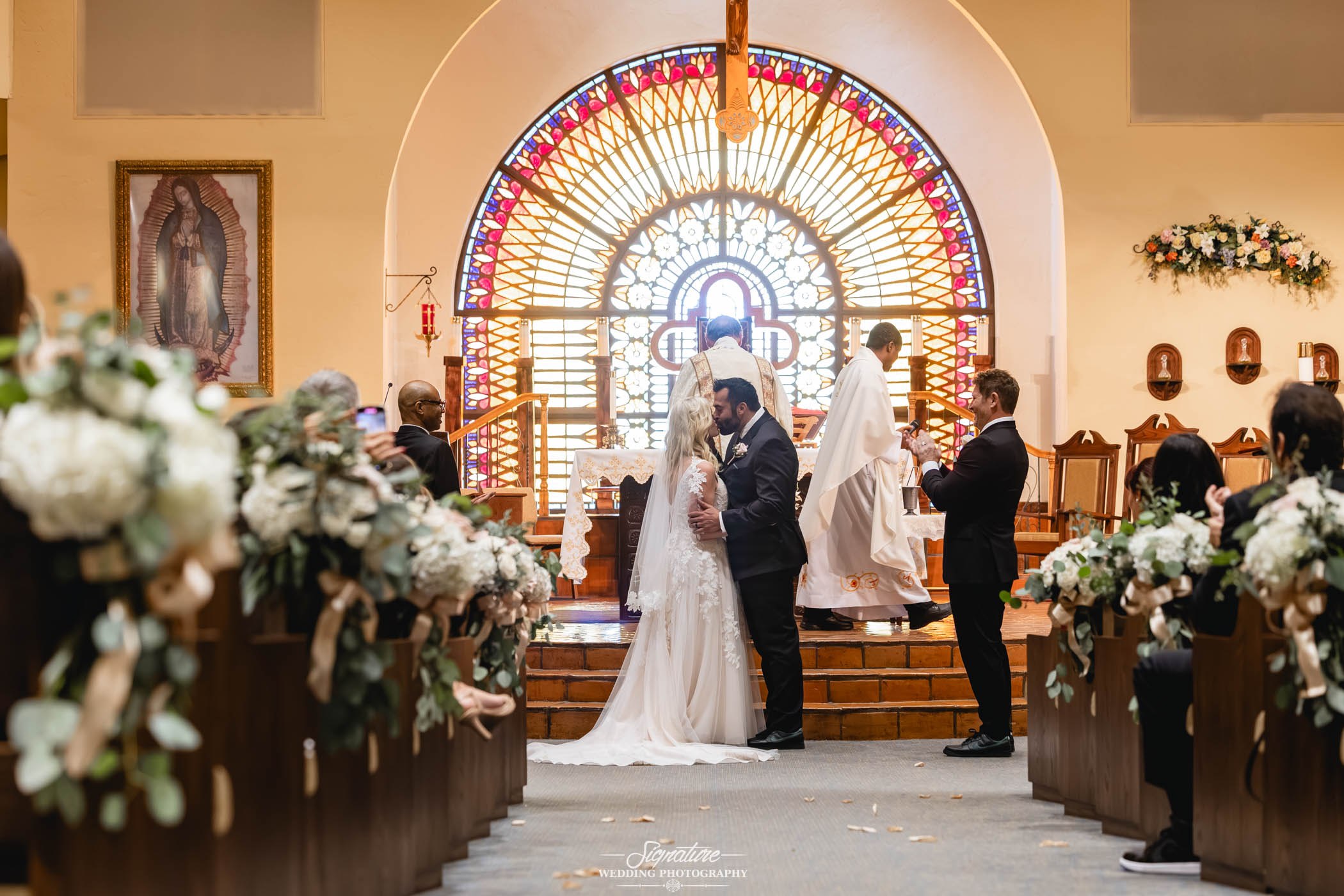 Image by Signature Wedding Photography A bride and groom kiss at the altar of a church, surrounded by floral decorations, guests seated in pews, and a priest standing nearby with a stained glass window in the background.
