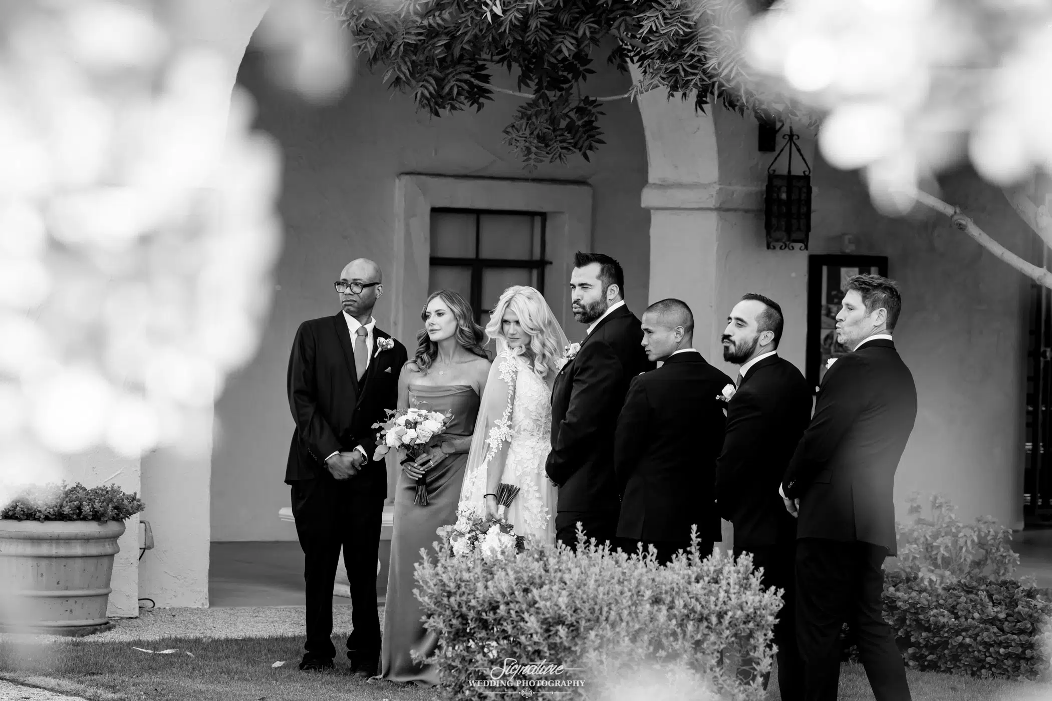 Image by Signature Wedding Photography A black and white photo of a bride and groom posing with their wedding party outdoors, standing near an arched building with greenery in the foreground. Everyone is dressed formally and looking toward the camera.