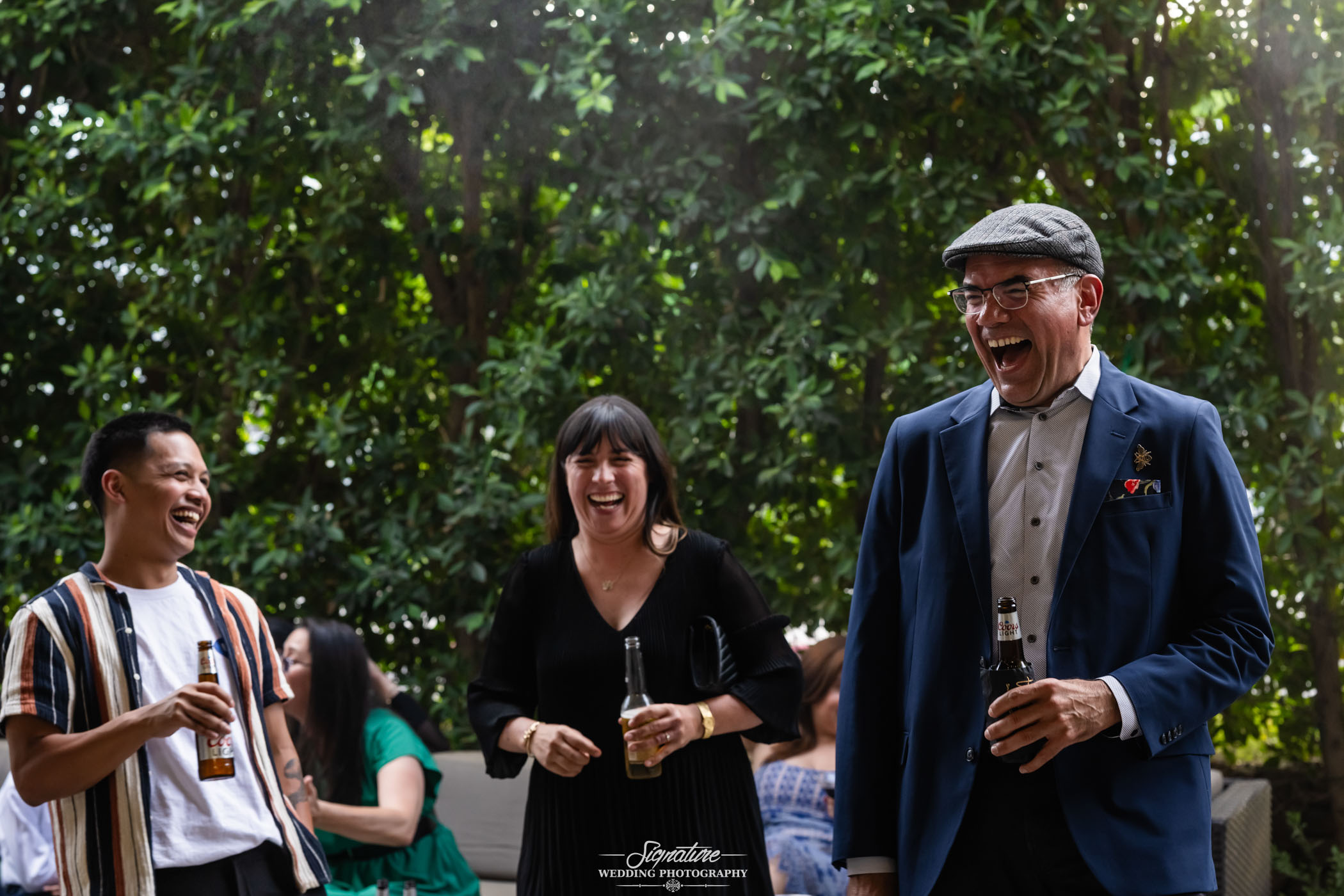 Image by Signature Wedding Photography Three people stand outdoors, laughing and holding drinks. A man in a blue suit and flat cap is on the right, a woman in a black dress is in the center, and a man in a striped shirt is on the left. Trees fill the background.