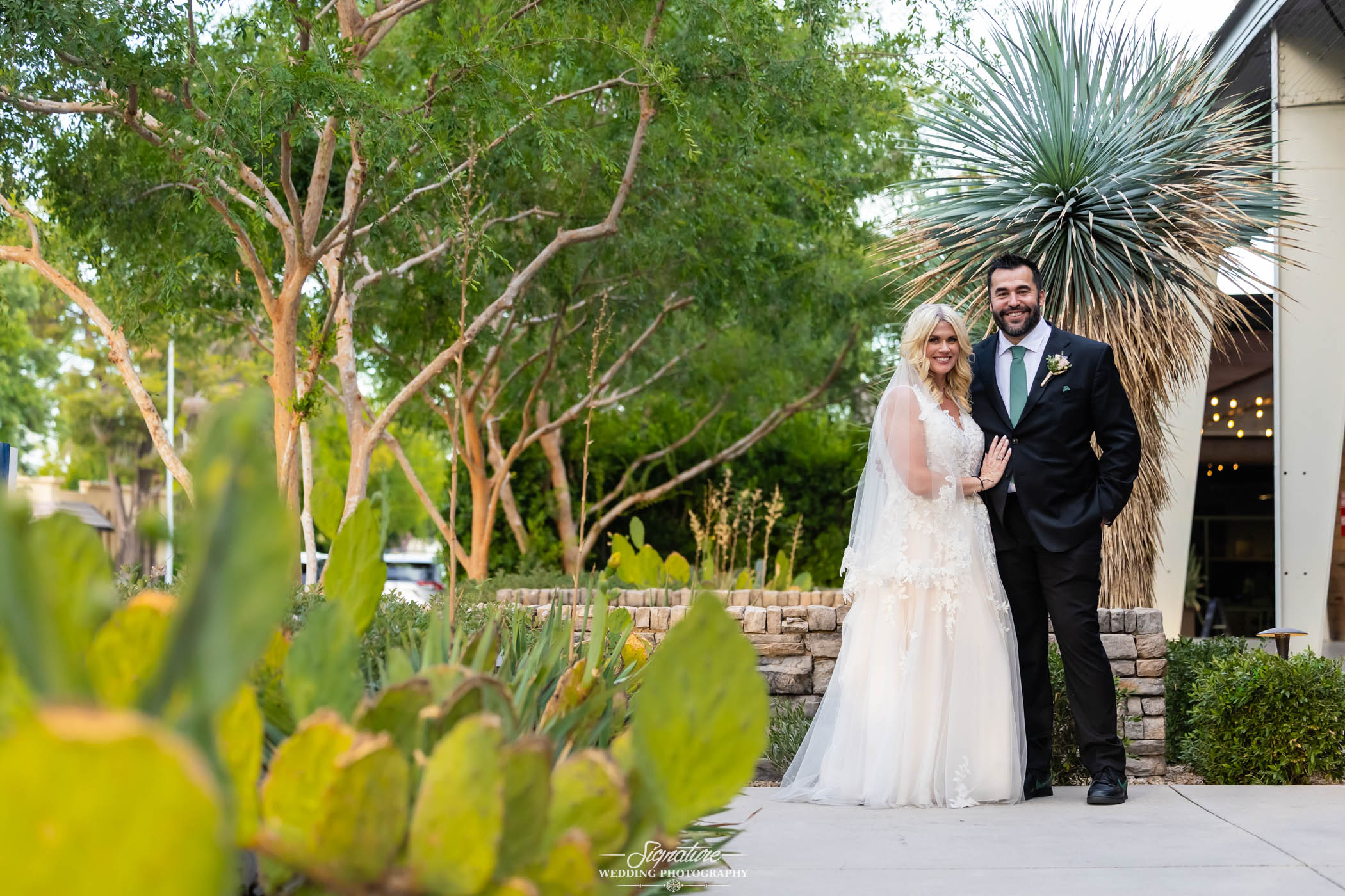 Image by Signature Wedding Photography A bride in a white gown and a groom in a black suit stand smiling together outdoors by a stone wall, surrounded by green trees and cacti.
