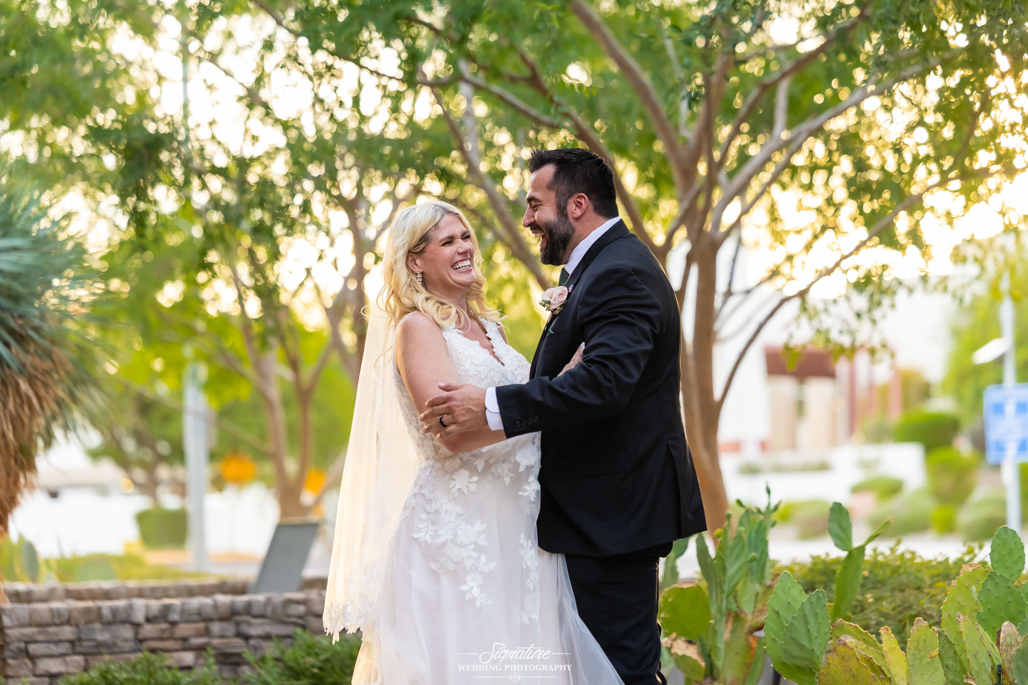 Image by Signature Wedding Photography A bride and groom stand outdoors in a garden, smiling and holding each other. The bride wears a white dress and veil, while the groom is in a black suit. Lush greenery and sunlight fill the background.