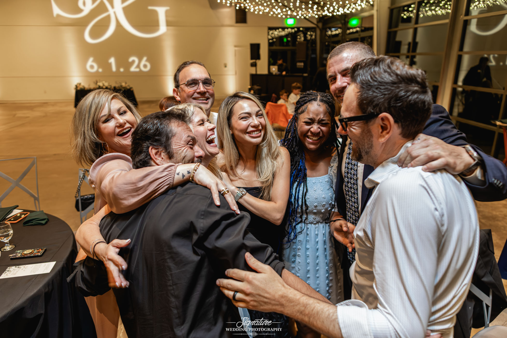 Image by Signature Wedding Photography A group of eight adults dressed in formal attire laugh and embrace each other in a bright, decorated event space with string lights overhead and large windows in the background.
