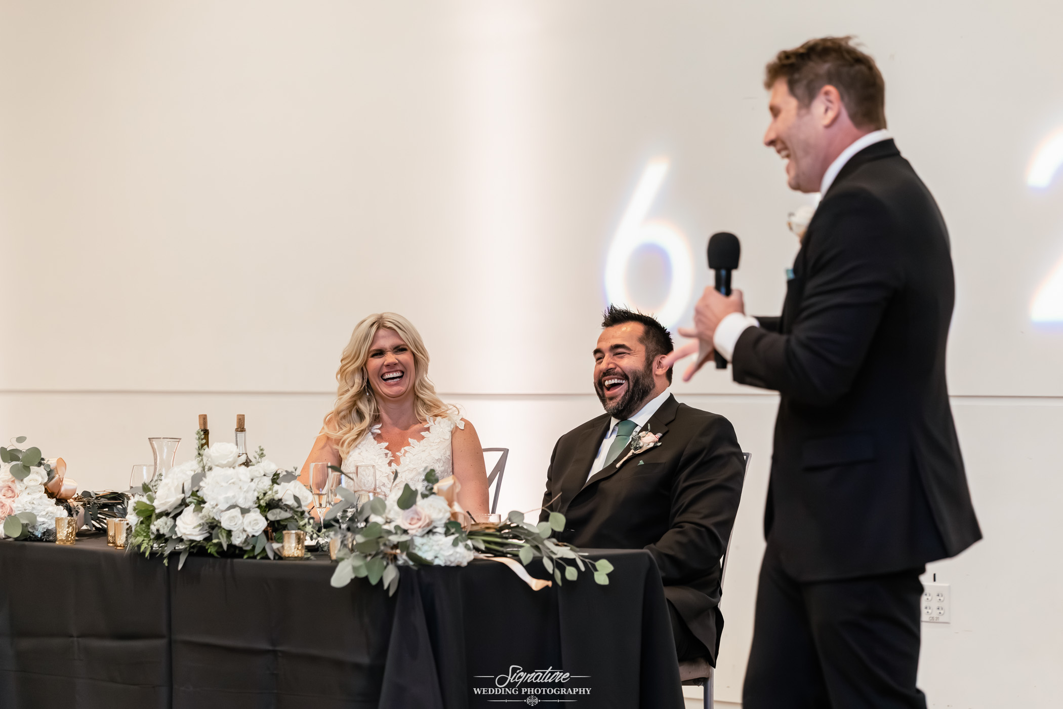 Image by Signature Wedding Photography A bride and groom sit at a decorated table, laughing joyfully as a man in a suit gives a speech with a microphone at a wedding reception. The setting is elegant with white flowers and candles.