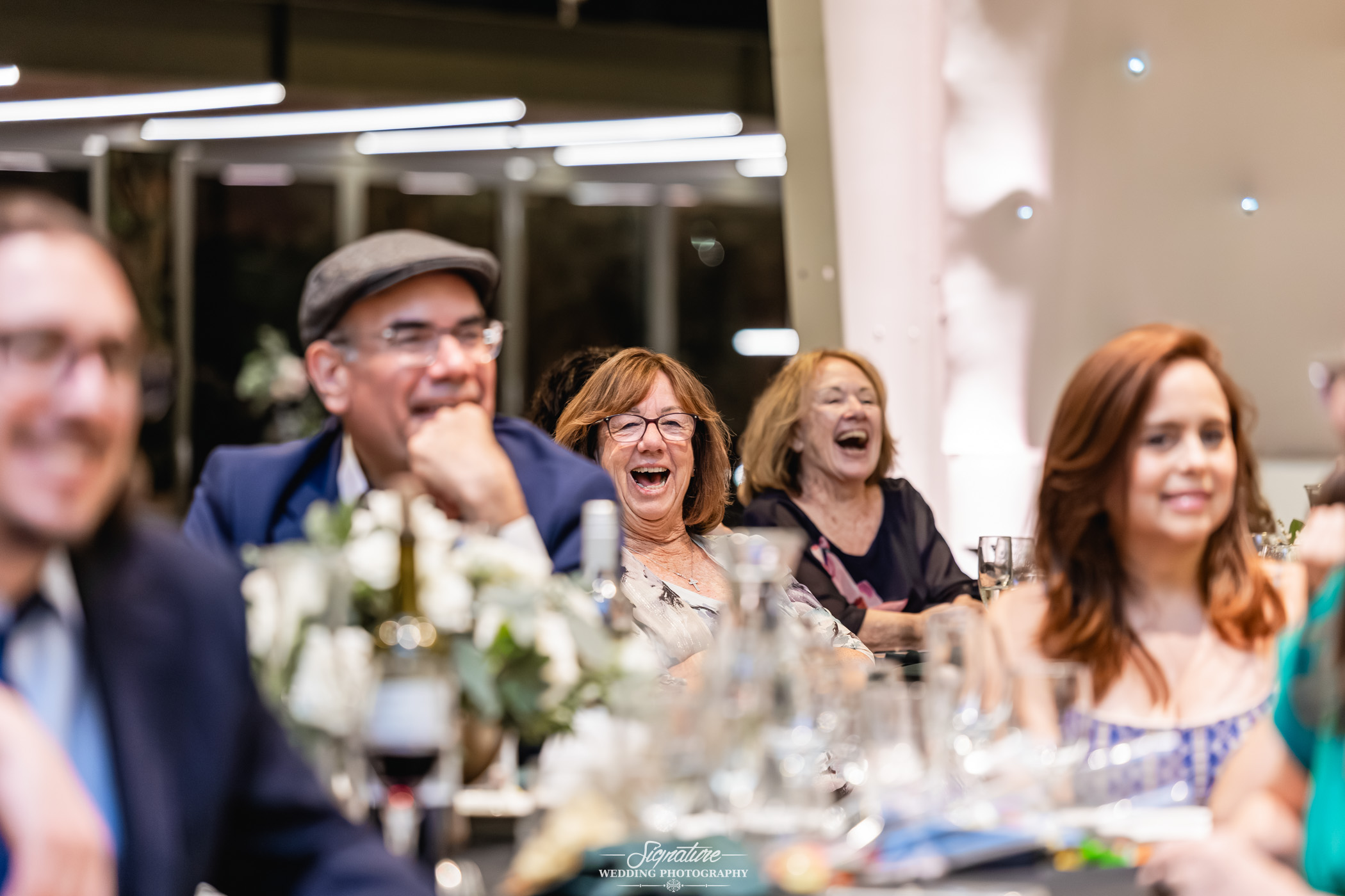 Image by Signature Wedding Photography A group of people sit at a decorated table, laughing and smiling during what appears to be a festive event or celebration. The background is softly lit, and the atmosphere is joyful and lively.