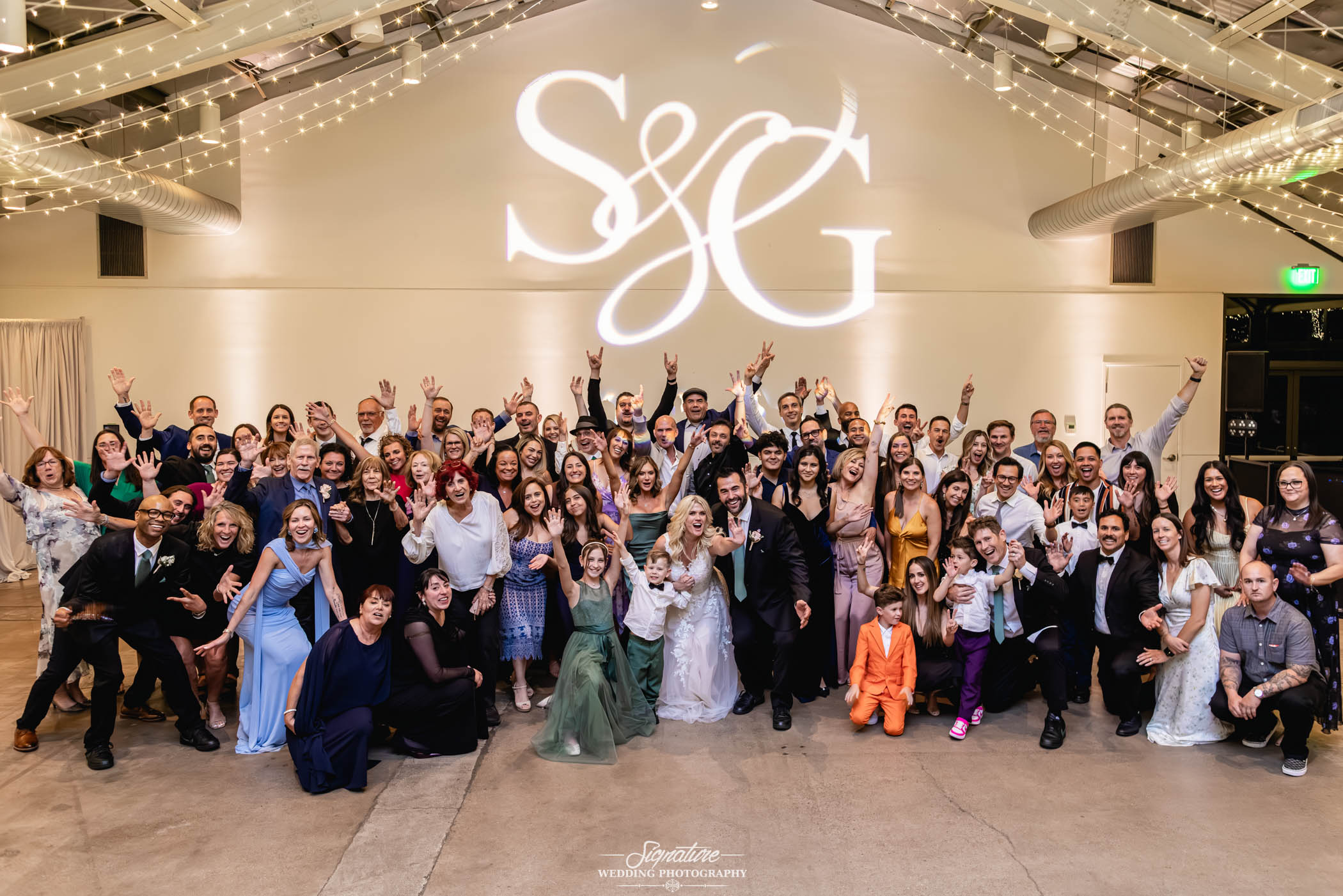 Image by Signature Wedding Photography A large group of wedding guests, including the bride and groom, pose together indoors under string lights, smiling and raising their arms in celebration. The initials "S & G" are projected on the wall behind them.