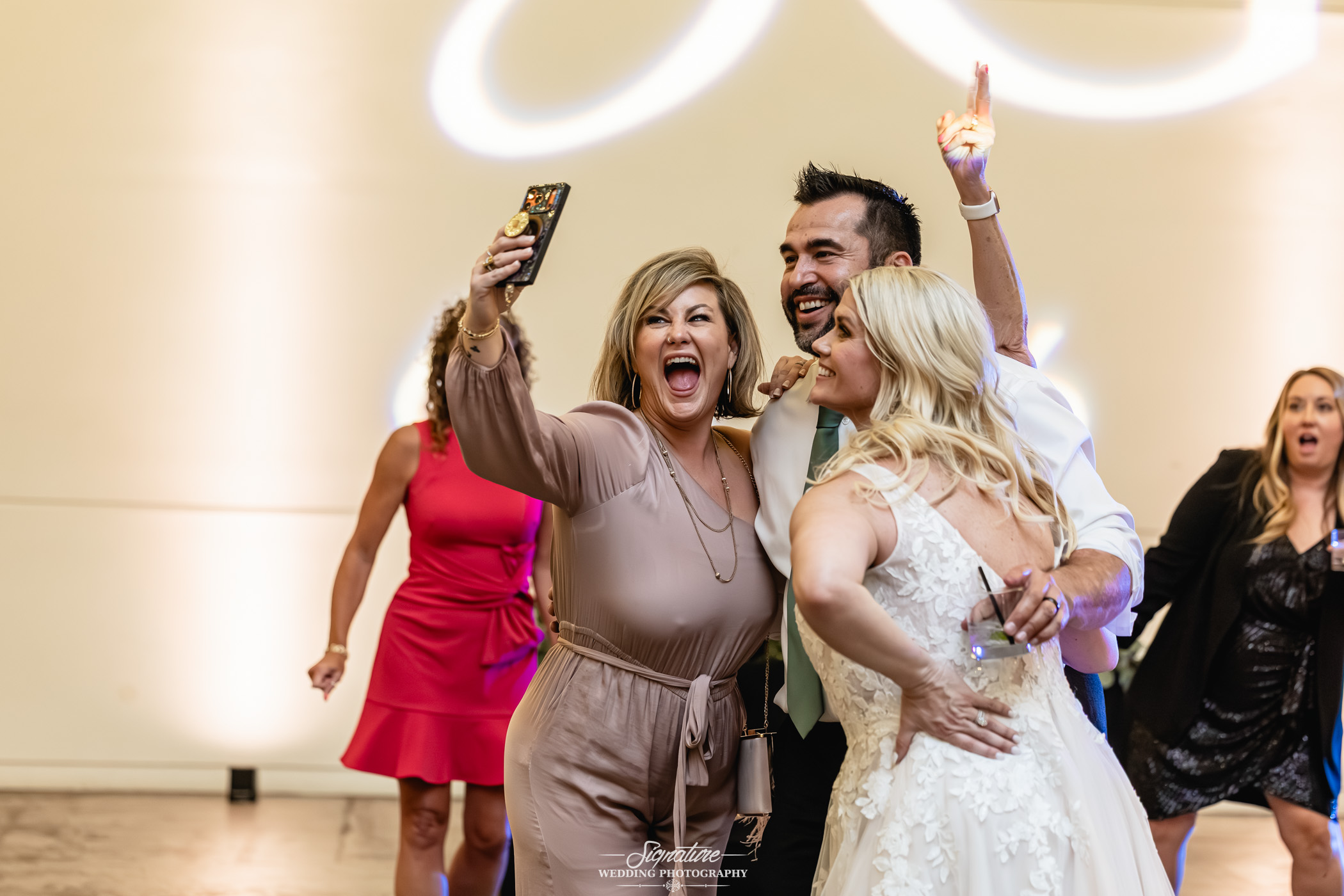 Image by Signature Wedding Photography A woman in a tan dress takes a joyful selfie with a smiling bride and groom on a dance floor, surrounded by guests celebrating at a wedding reception.