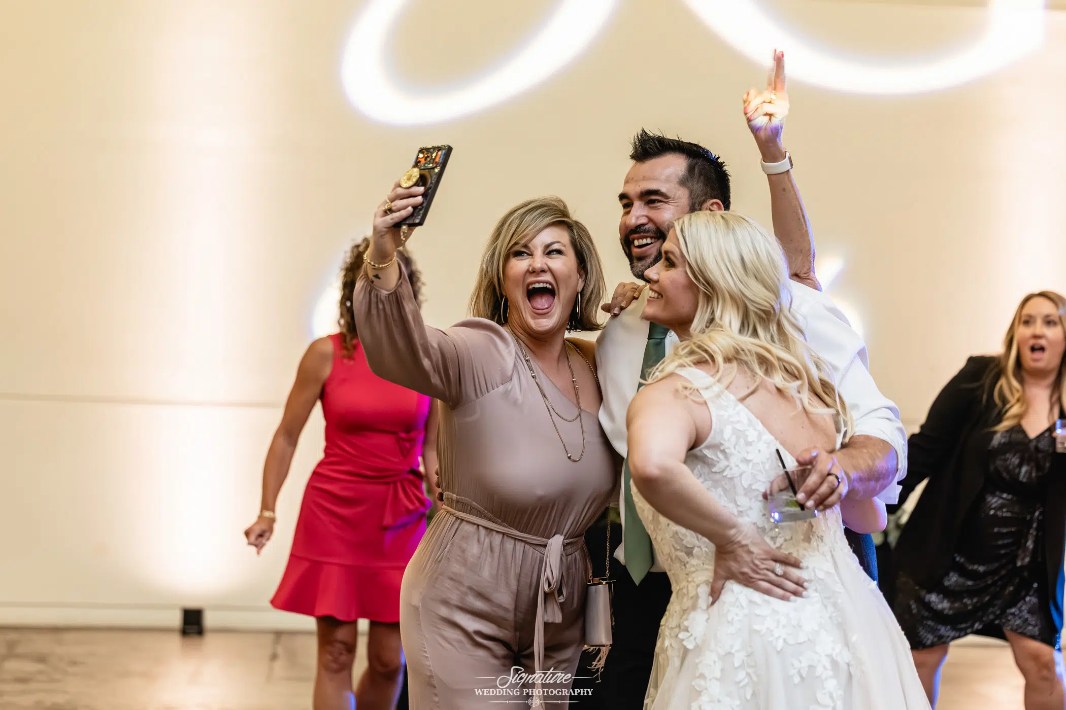 Image by Signature Wedding Photography A woman in a tan dress takes a joyful selfie with a smiling bride and groom on a dance floor, surrounded by guests celebrating at a wedding reception.