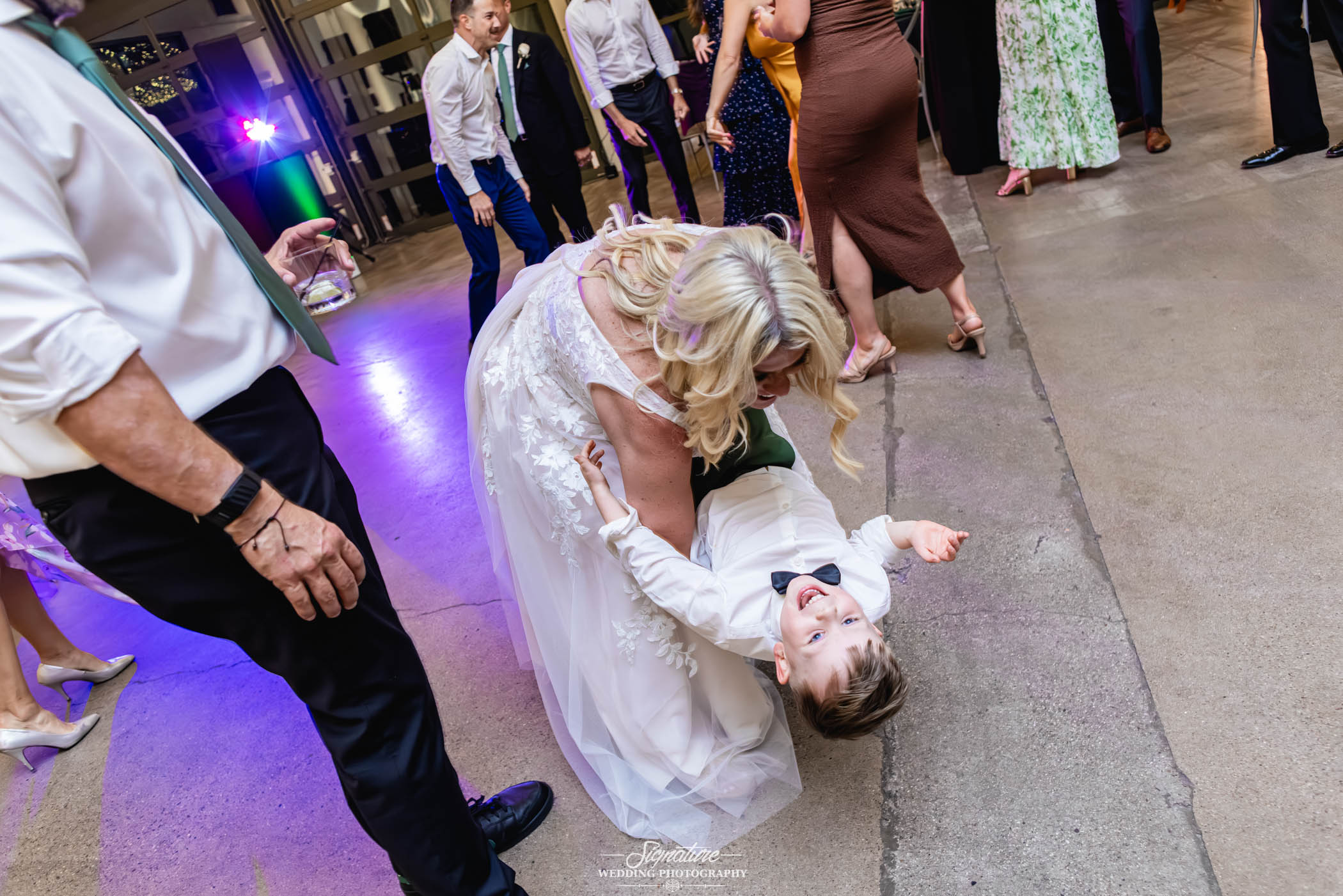 Image by Signature Wedding Photography A woman in a white dress playfully dips a laughing young boy in dress clothes on the dance floor at a lively indoor event, surrounded by other dancing guests.