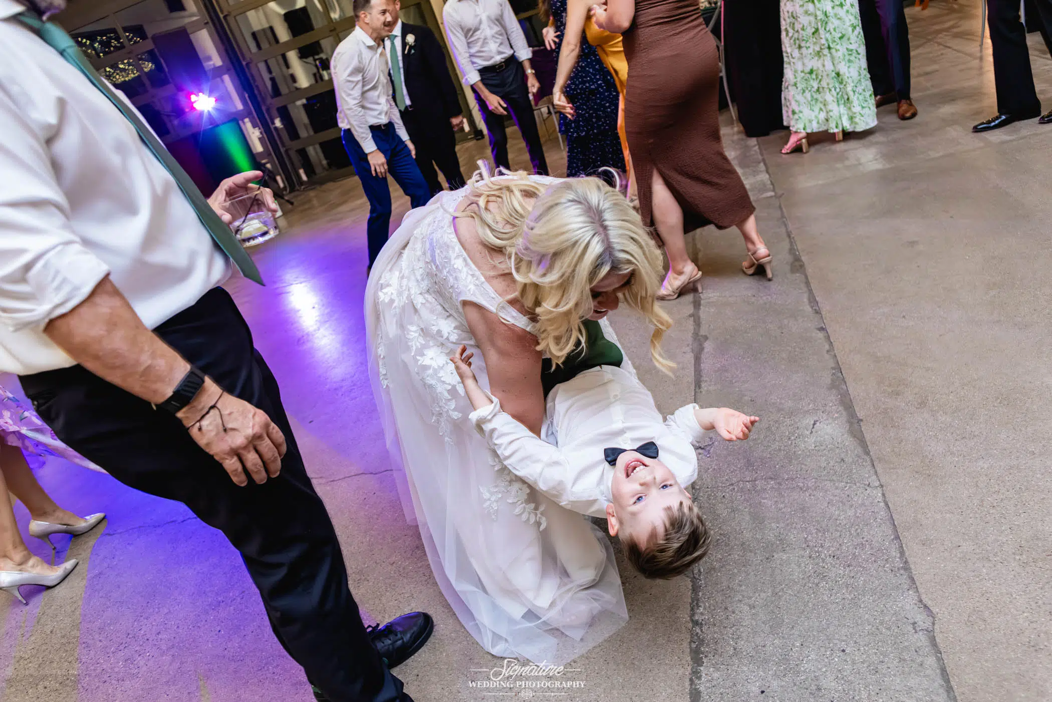 Image by Signature Wedding Photography A woman in a white dress playfully dips a laughing young boy in dress clothes on the dance floor at a lively indoor event, surrounded by other dancing guests.