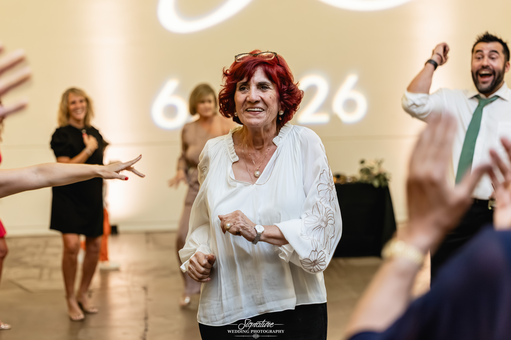 Image by Signature Wedding Photography A joyful older woman with red hair and a white blouse dances at an indoor event, surrounded by other smiling people who are also dancing and enjoying the celebration.