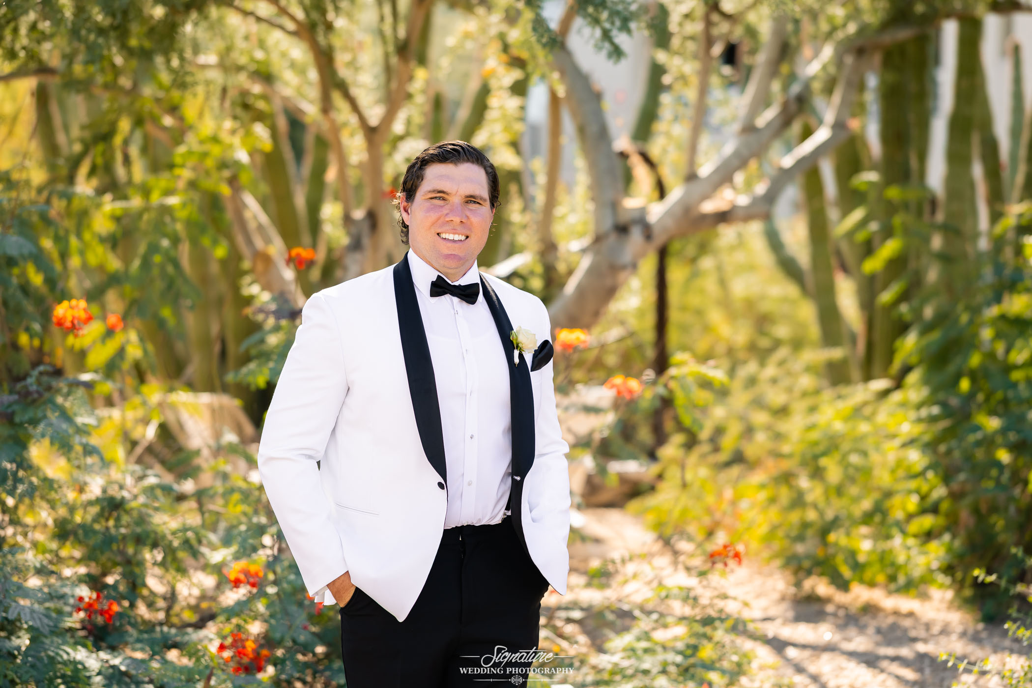 Image by Signature Wedding Photography A man wearing a white tuxedo jacket, black bow tie, and black pants stands outdoors in a garden with green foliage and orange flowers, smiling at the camera.