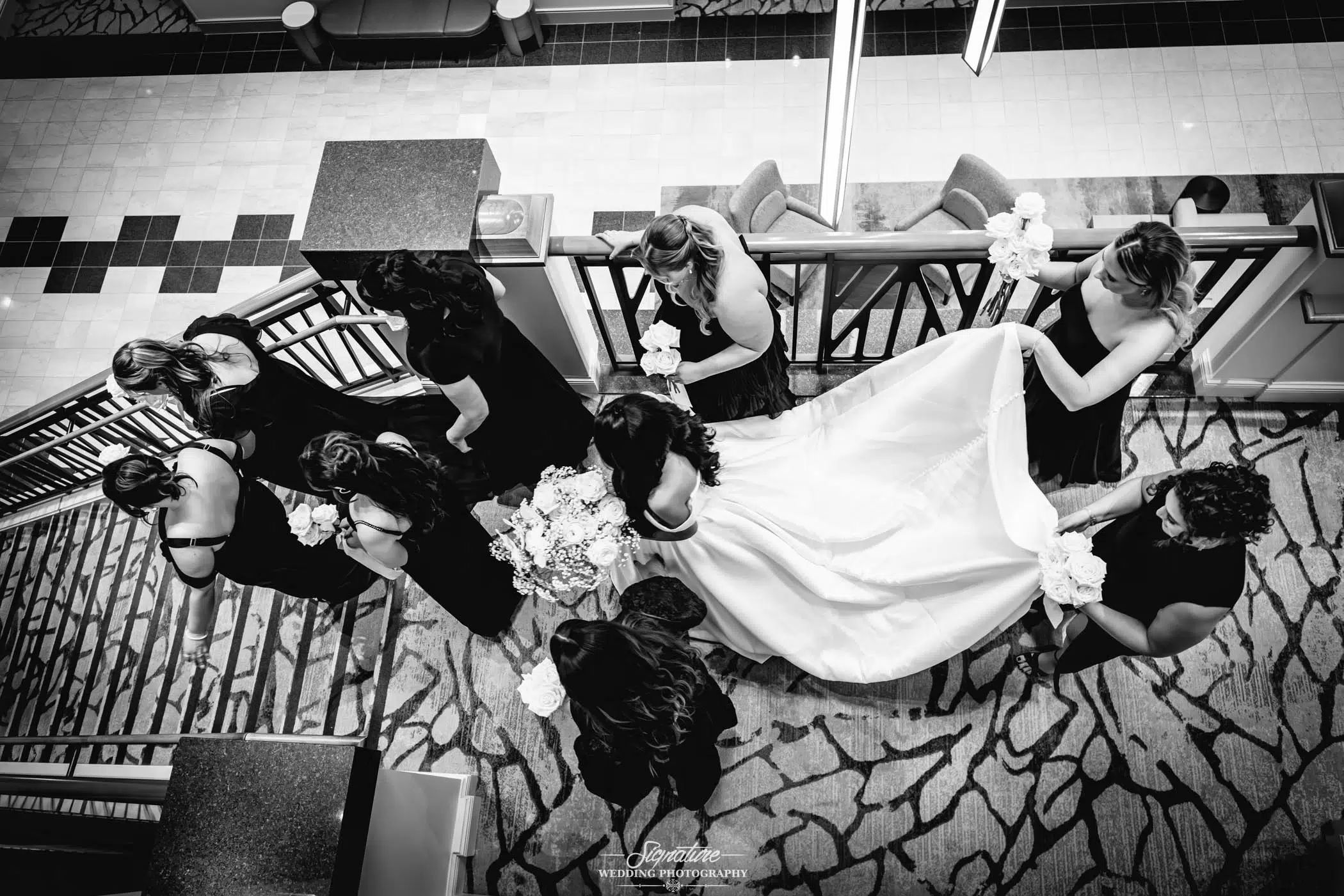 Image by Signature Wedding Photography Black and white overhead photo of a bride holding a bouquet, surrounded by bridesmaids in dark dresses who help arrange her long gown on a patterned carpeted floor. The group stands near a staircase railing.