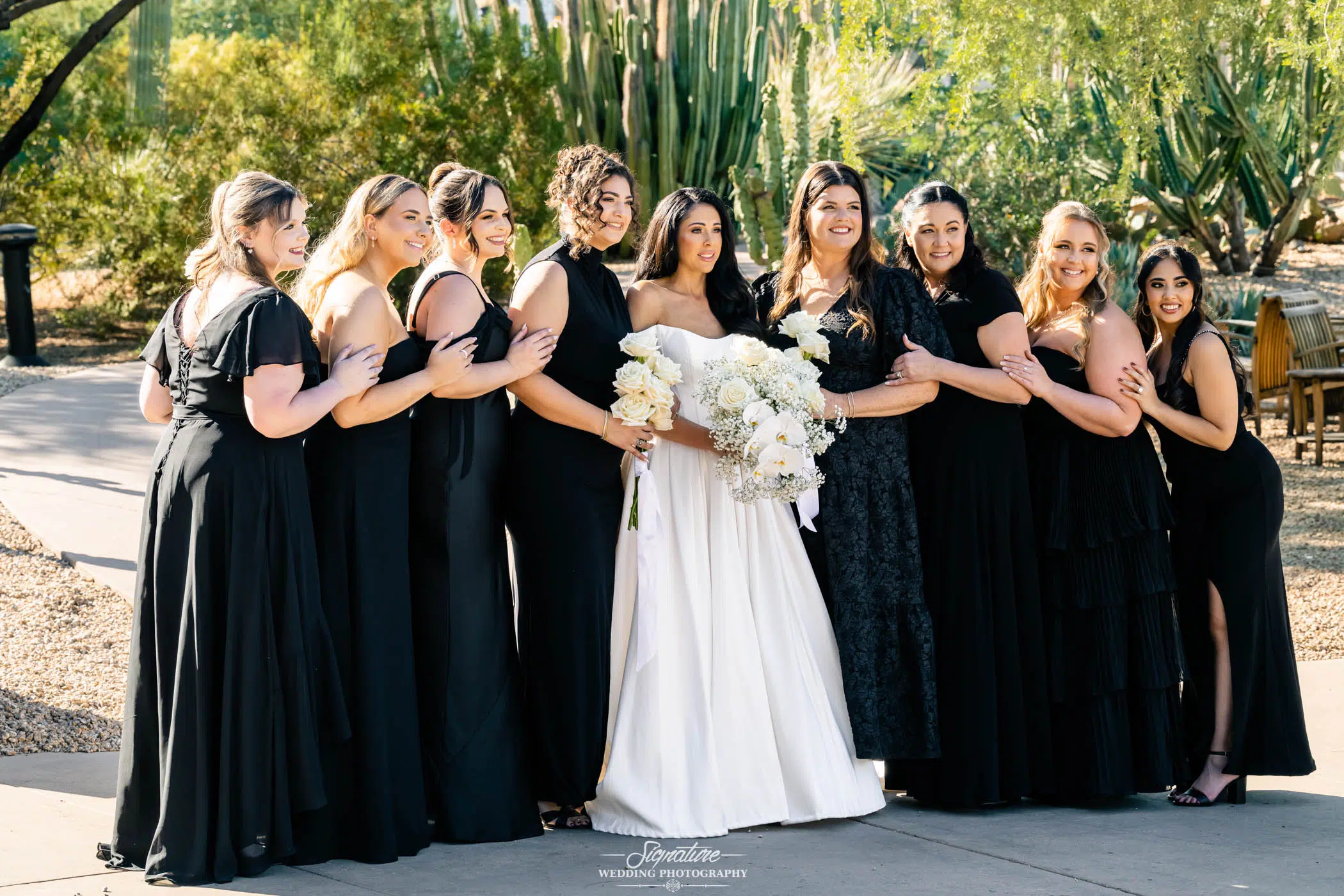 Image by Signature Wedding Photography A bride in a white dress holding a bouquet of white flowers stands outdoors, surrounded by eight bridesmaids in black dresses, all smiling and posing together in a garden setting with cacti and greenery.