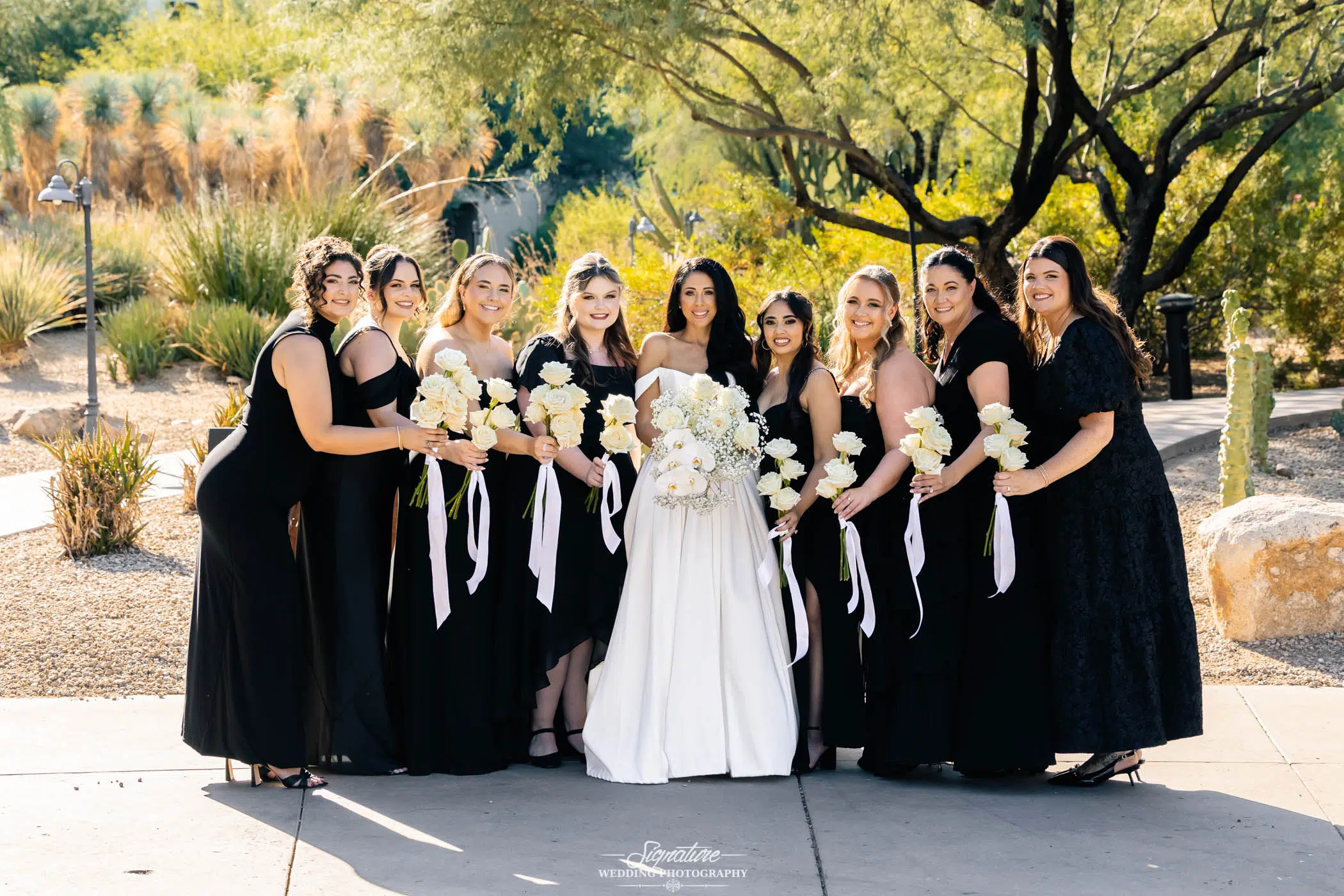 Image by Signature Wedding Photography A bride in a white gown stands outdoors with eight bridesmaids in black dresses, all holding white bouquets and smiling at the camera against a backdrop of trees and desert plants.