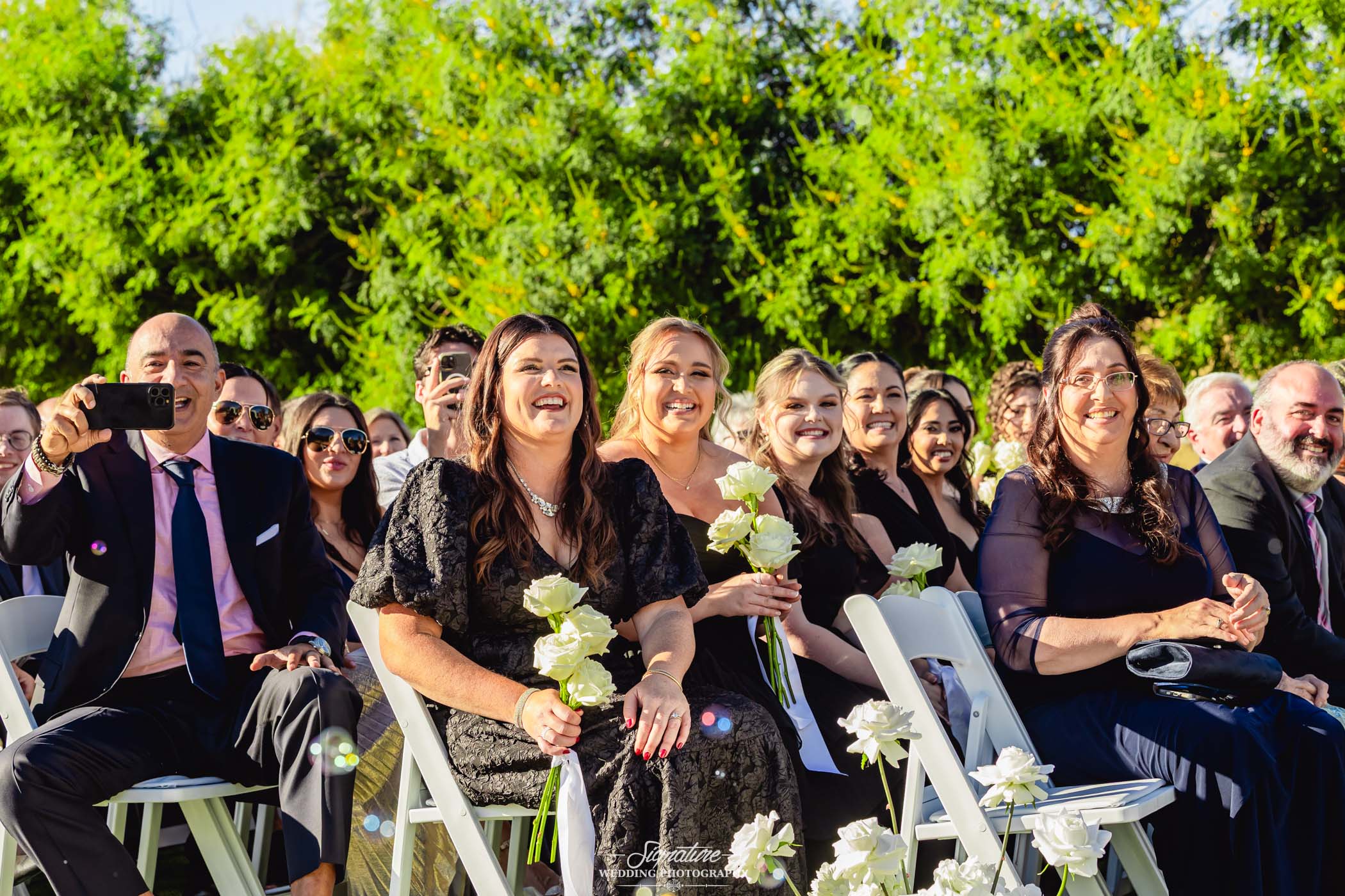 Image by Signature Wedding Photography A group of well-dressed wedding guests sit outdoors on white chairs, smiling and holding white roses. Some are taking photos, and bright sunlight highlights the joyful atmosphere, with greenery in the background.