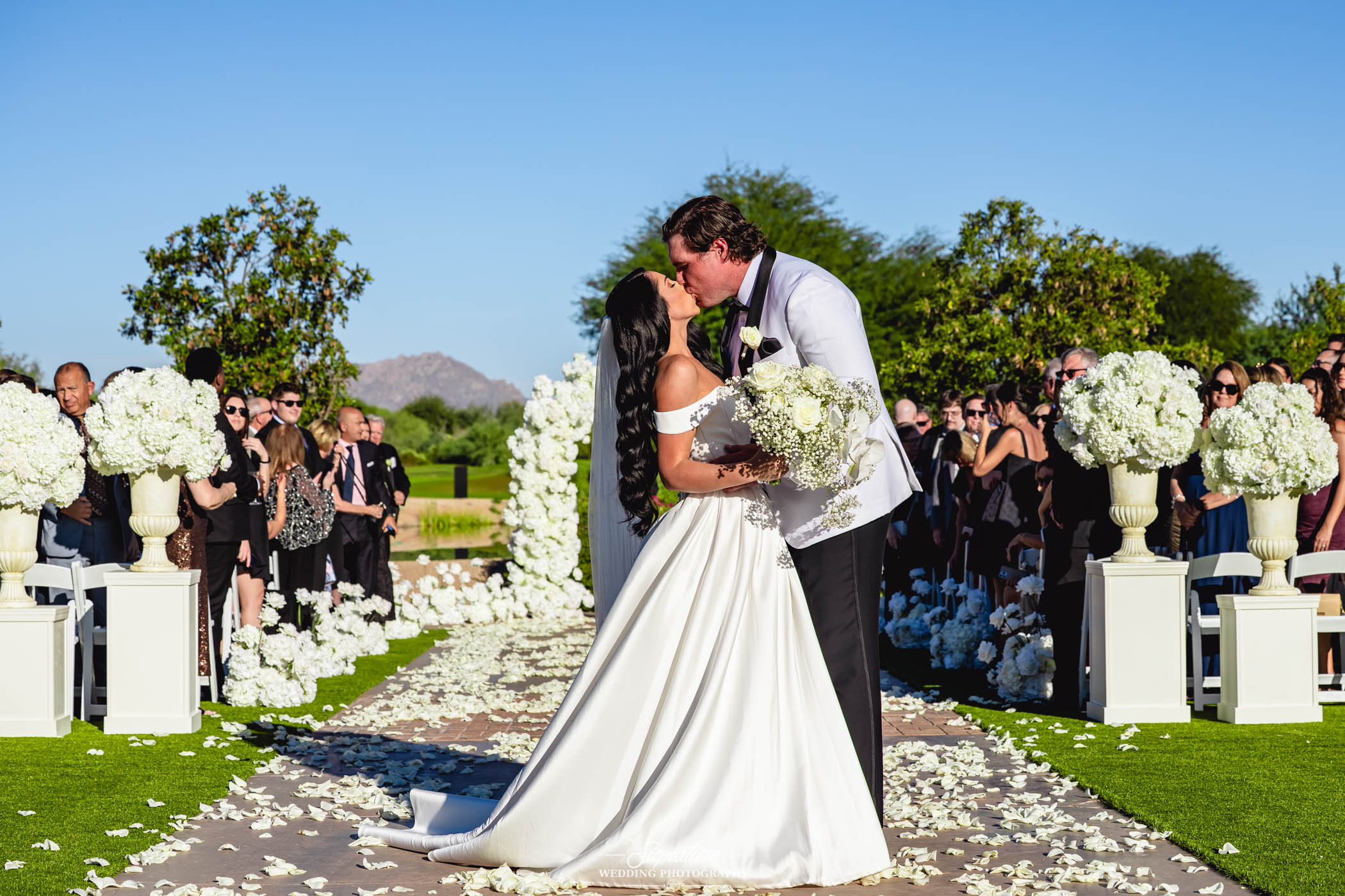 Image by Signature Wedding Photography A bride and groom kiss at an outdoor wedding ceremony, surrounded by white flowers and guests seated on either side of the aisle under a clear blue sky.
