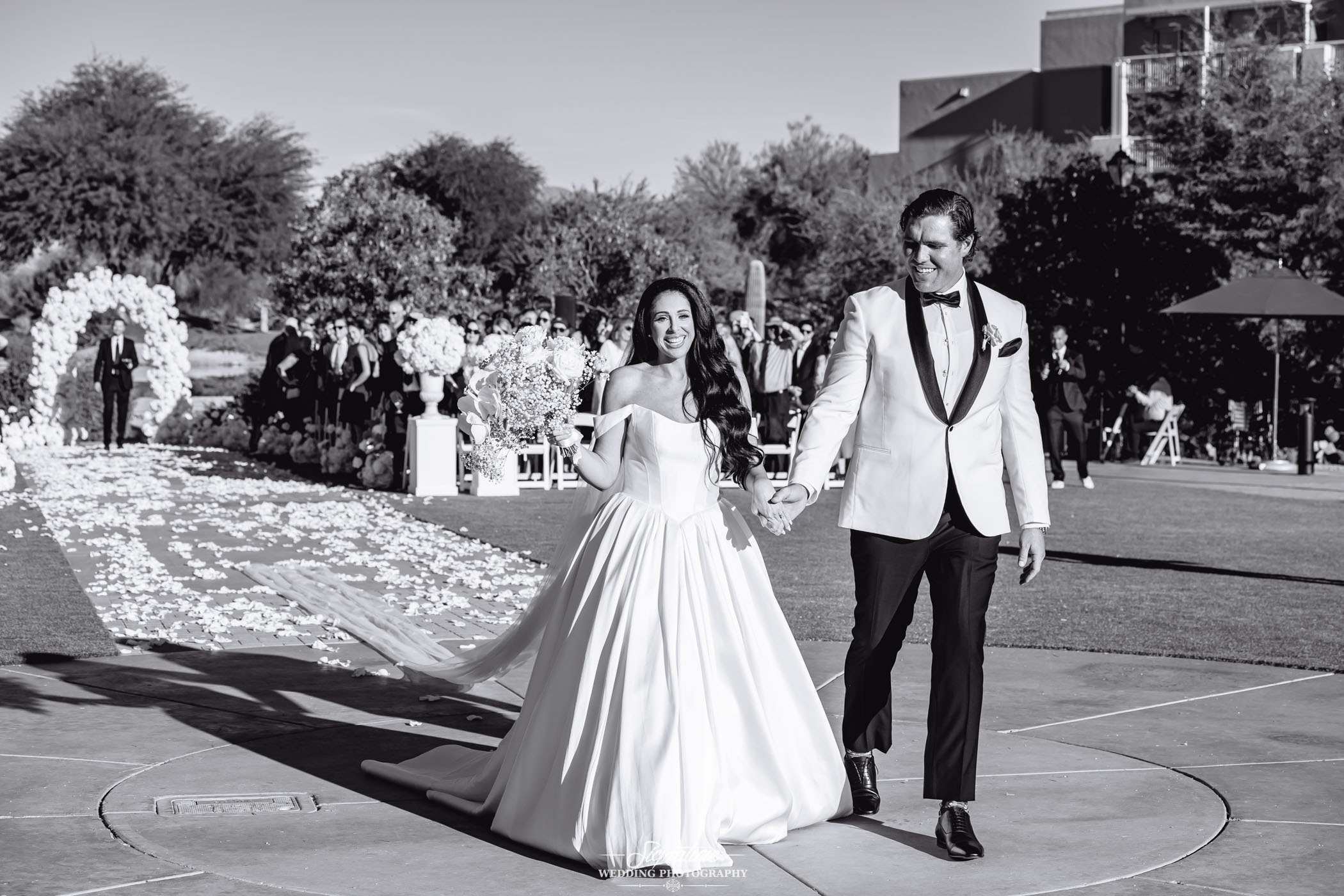 Image by Signature Wedding Photography A bride in a flowing white gown and a groom in a white tuxedo jacket hold hands and smile as they walk down an outdoor aisle lined with flower petals, with guests and floral arrangements in the background.
