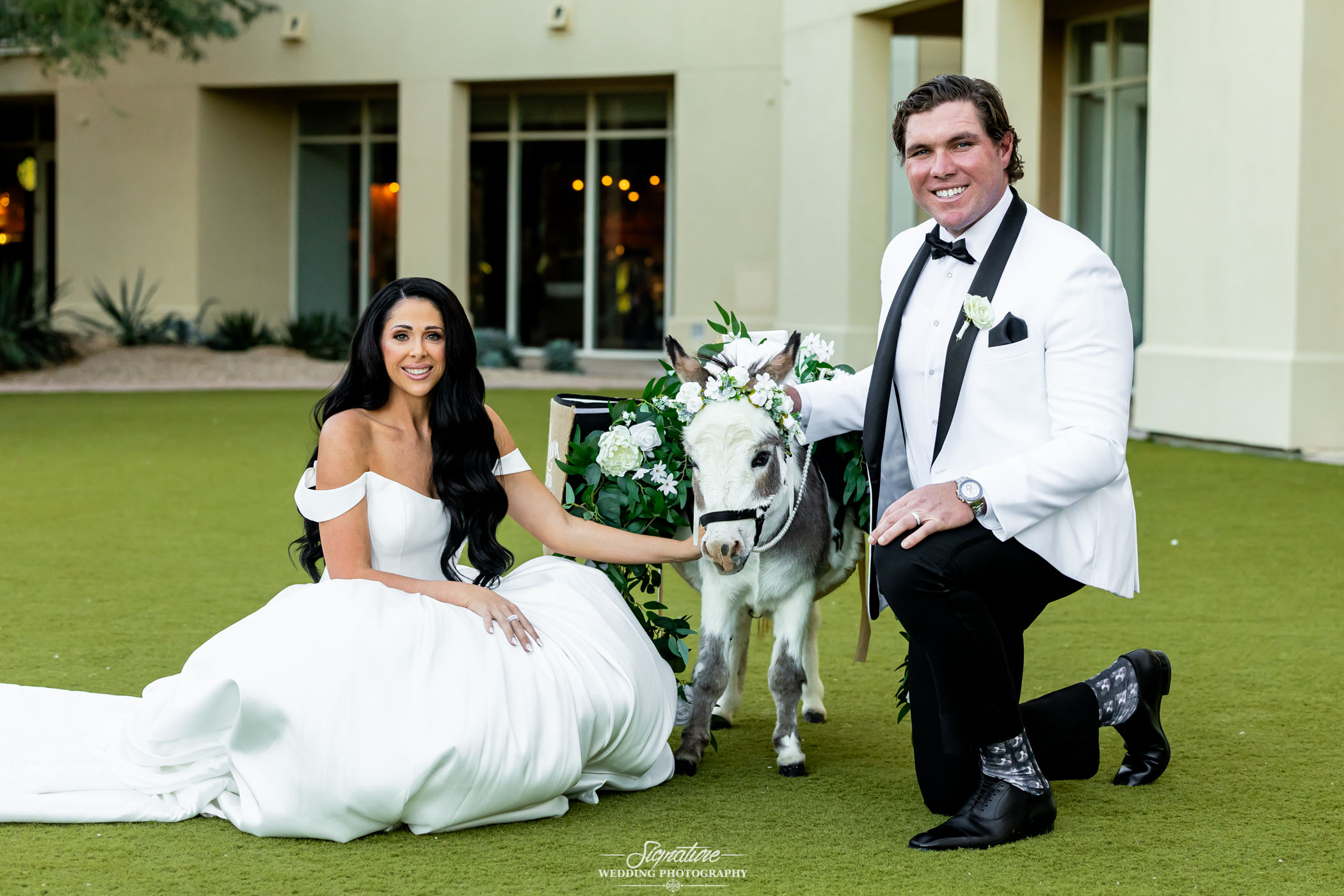 Image by Signature Wedding Photography A bride in a white gown and a groom in a white tuxedo jacket kneel on grass beside a small donkey adorned with greenery and white flowers, outside a building.
