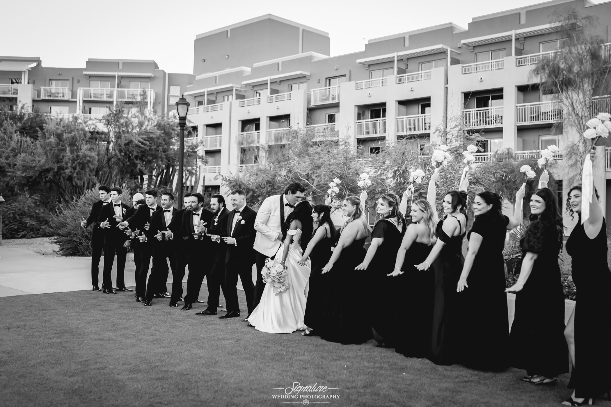 Image by Signature Wedding Photography A wedding party poses outdoors in front of a modern building; the bride and groom stand in the center, surrounded by bridesmaids in black dresses and groomsmen in tuxedos, all smiling and holding bouquets.