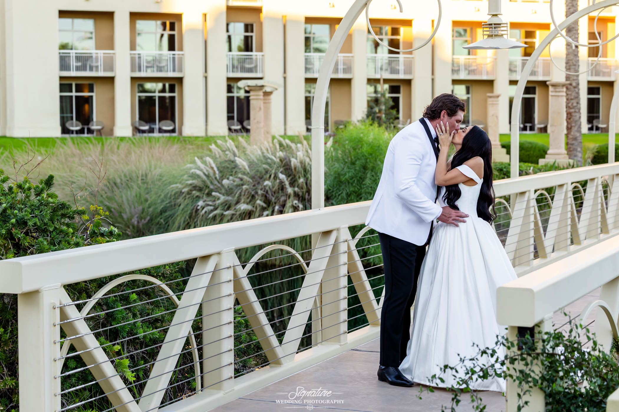 Image by Signature Wedding Photography A bride in a white gown and a groom in a white tuxedo jacket share a kiss on a bridge outside a building with balconies and lush greenery.