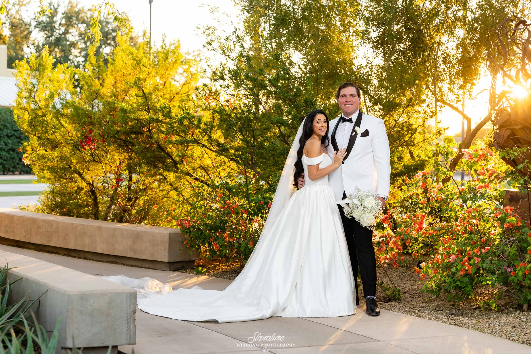 Image by Signature Wedding Photography A bride in a white gown and a groom in a white tuxedo jacket stand together outdoors among green and flowering bushes at sunset, smiling and holding a bouquet.