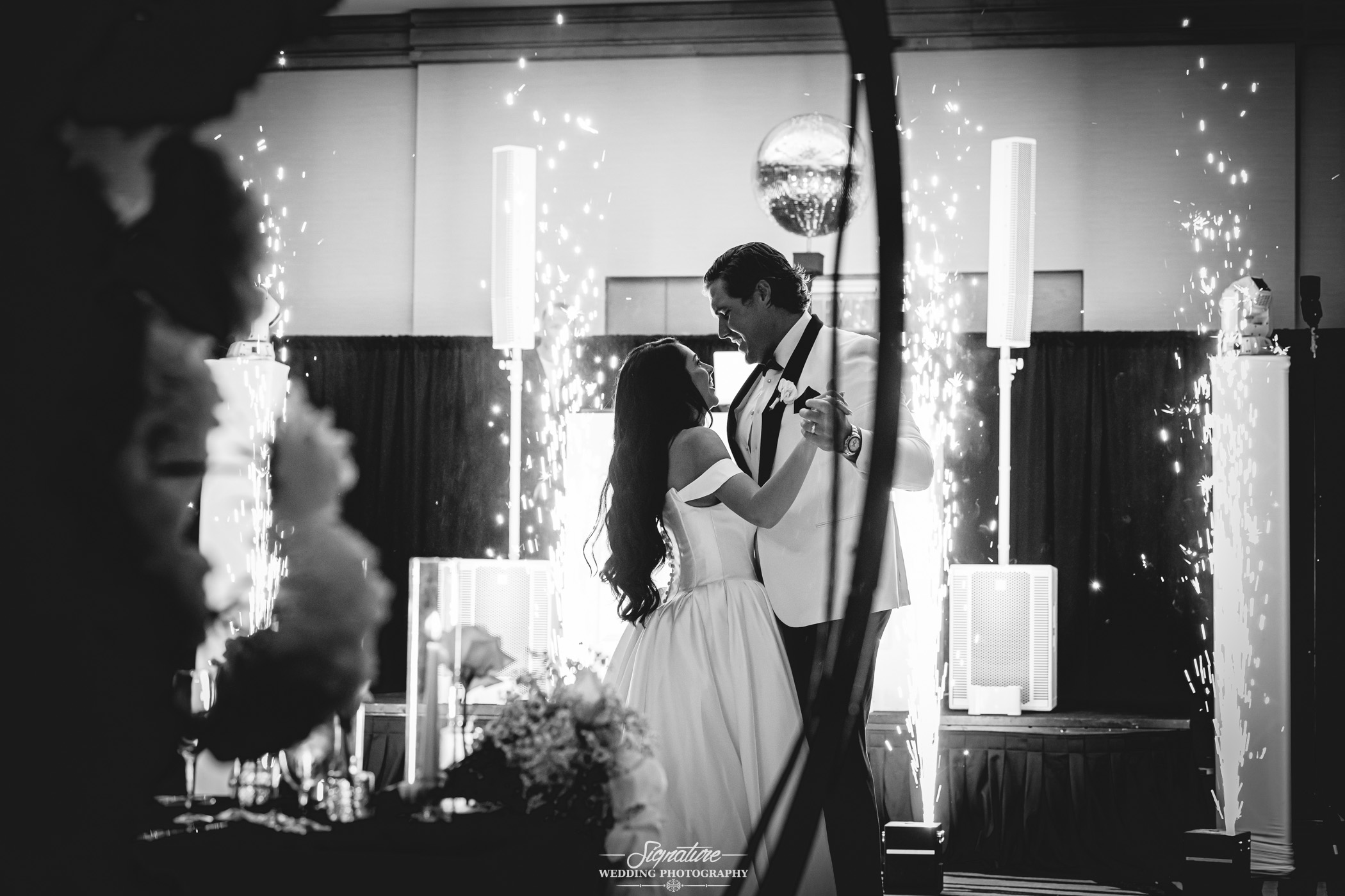Image by Signature Wedding Photography A bride and groom share a first dance under sparkling fireworks indoors, surrounded by elegant decor, in a black-and-white photo capturing a joyful wedding moment.