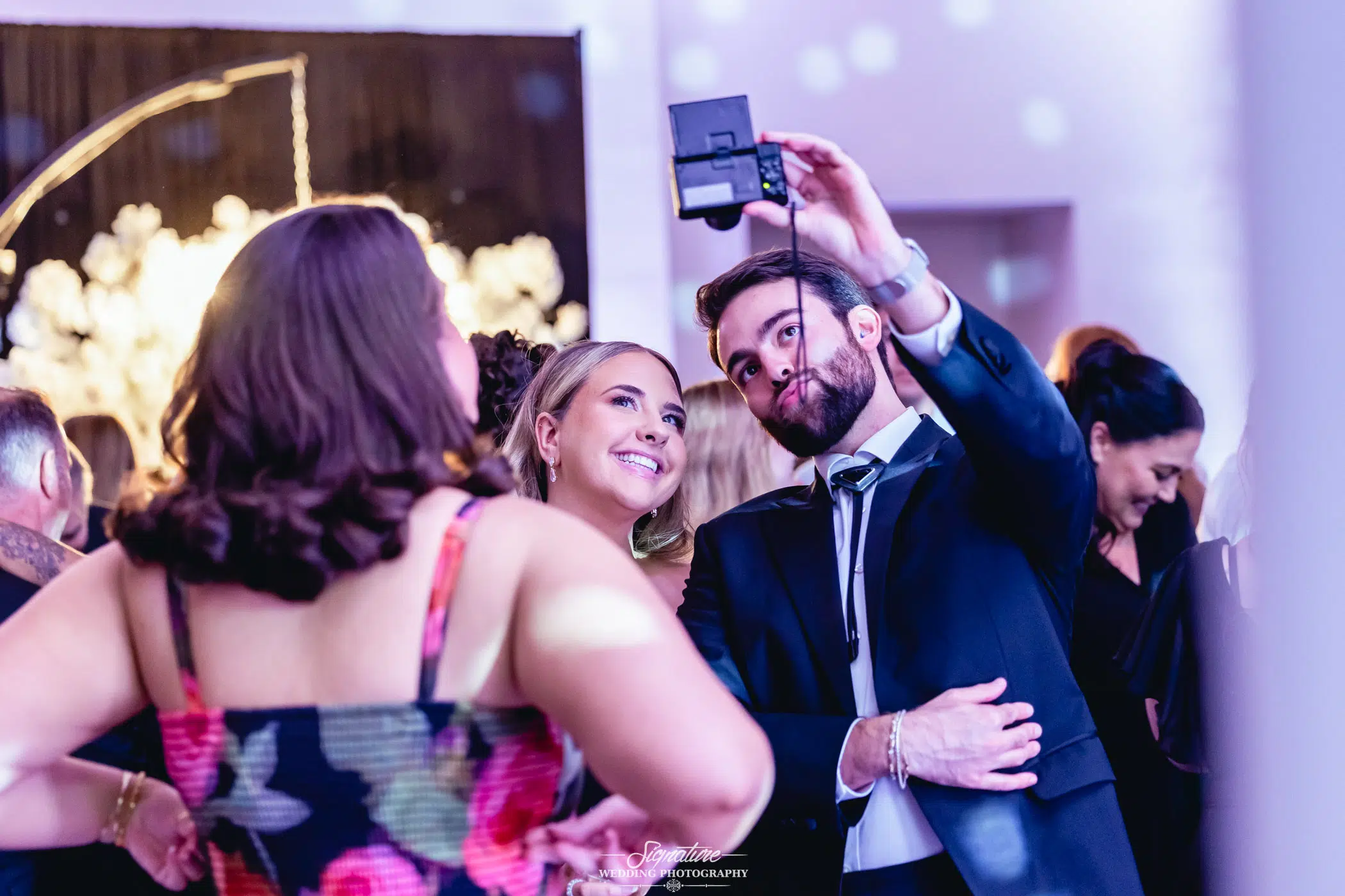 Image by Signature Wedding Photography A man in a suit holds up a phone to take a selfie with a smiling woman in a floral dress at a lively indoor event, surrounded by other people in formal attire.