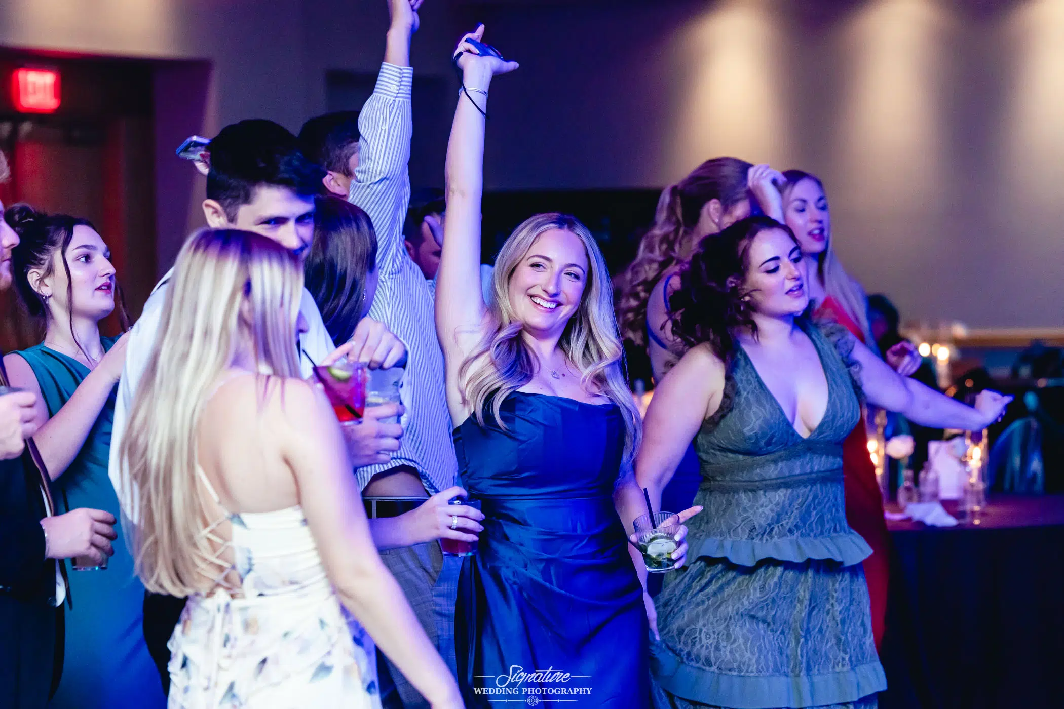 Image by Signature Wedding Photography A group of people dressed in formal attire dance and celebrate at an indoor event. A woman in a blue dress stands in the center, smiling with one arm raised, holding a drink. The atmosphere is lively and joyful.