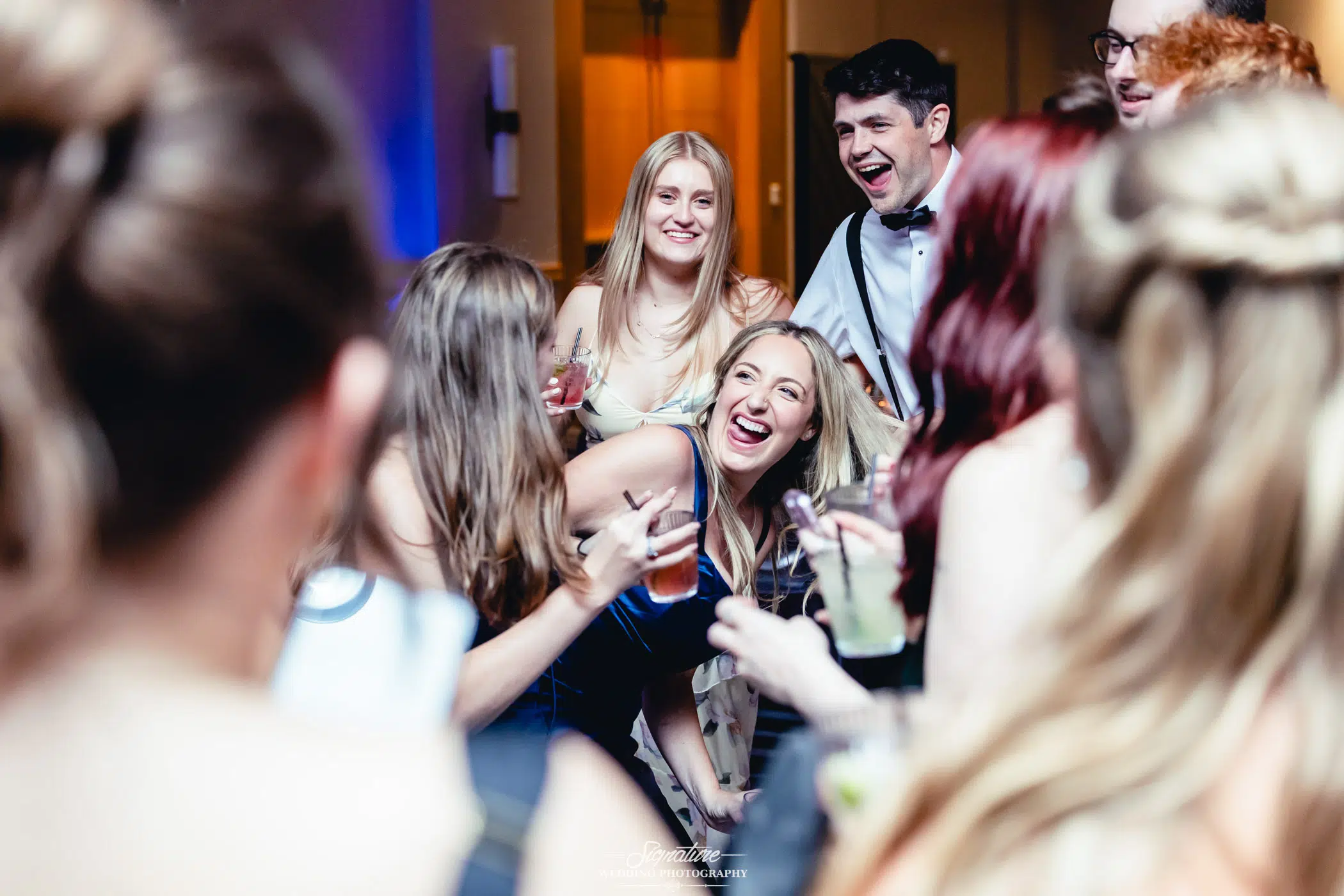 Image by Signature Wedding Photography A group of people dressed formally laugh and enjoy drinks together at an indoor event, with one woman in the center bending forward and smiling brightly. The scene is lively and joyful.