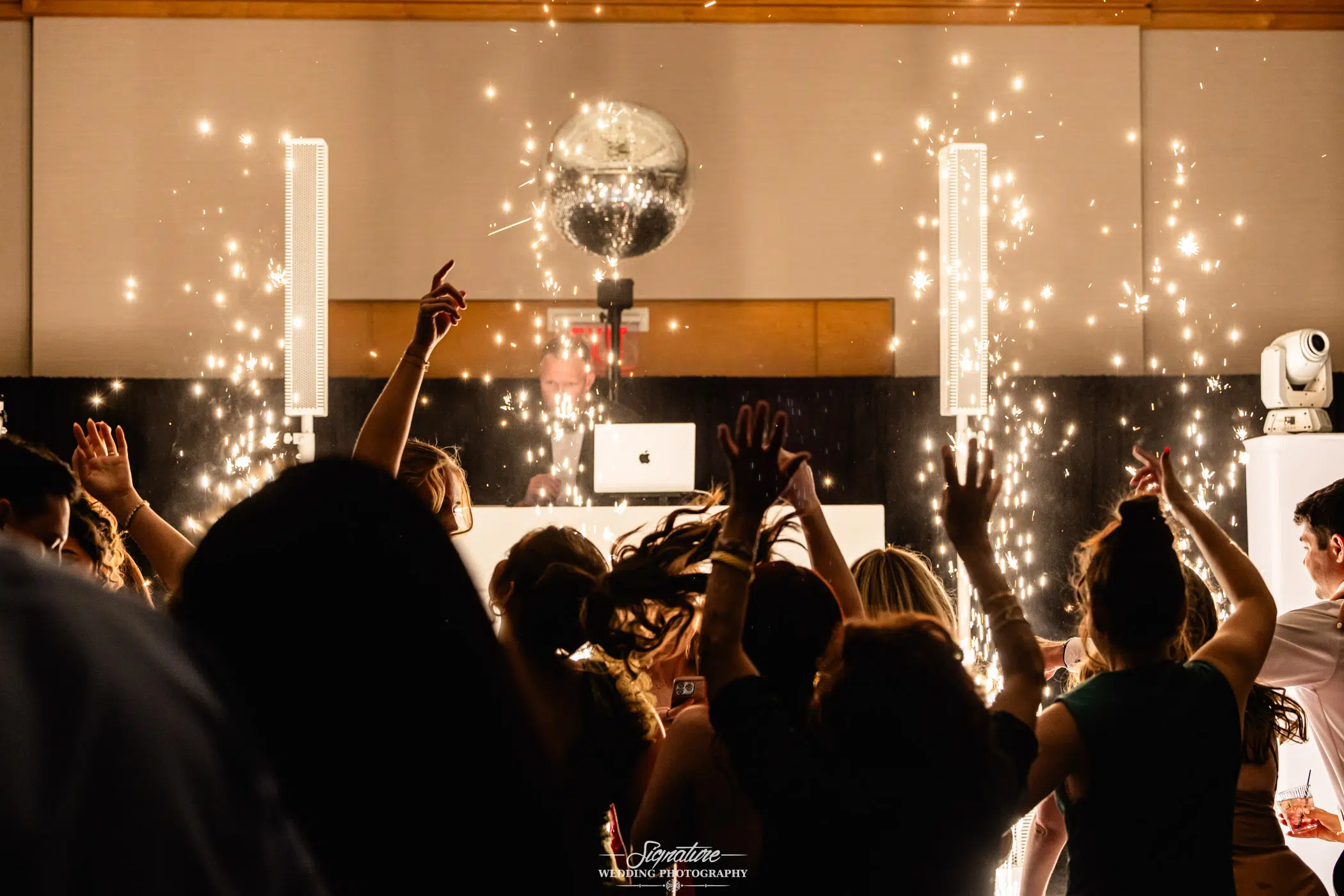 Image by Signature Wedding Photography A crowd of people dance with raised hands in front of a DJ booth, surrounded by bright sparklers and lights, with a disco ball reflecting above the lively scene.