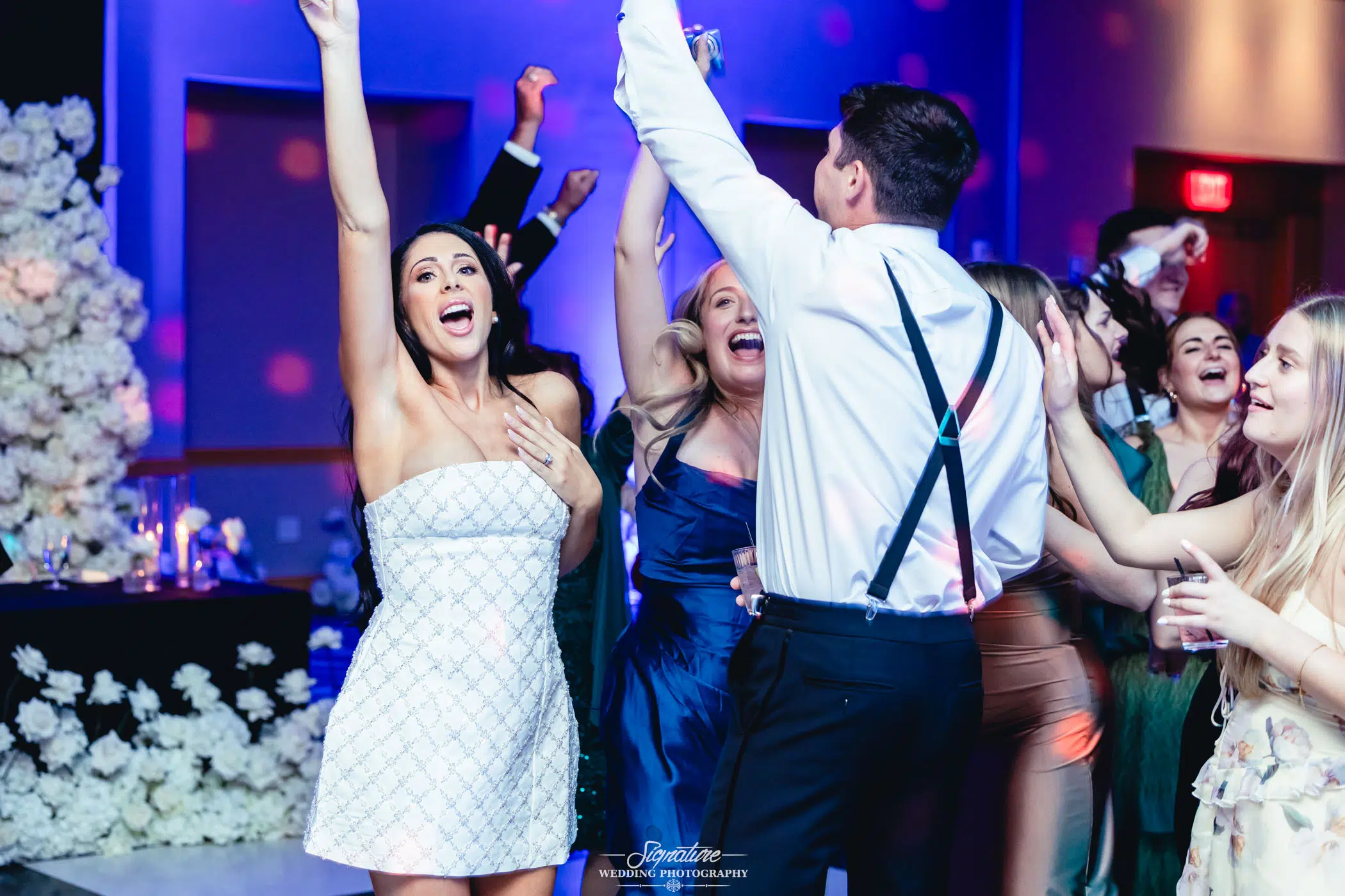 Image by Signature Wedding Photography A group of well-dressed people are dancing and cheering joyfully at a lively indoor event, with colorful lighting and floral decorations in the background.
