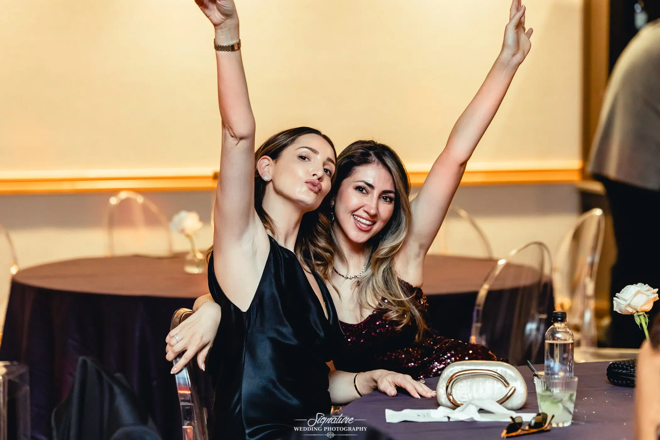 Image by Signature Wedding Photography Two women sitting at a table, smiling and raising their arms in celebration. The table has a water bottle, a clutch, and some flowers. The background shows empty chairs and a festive atmosphere.