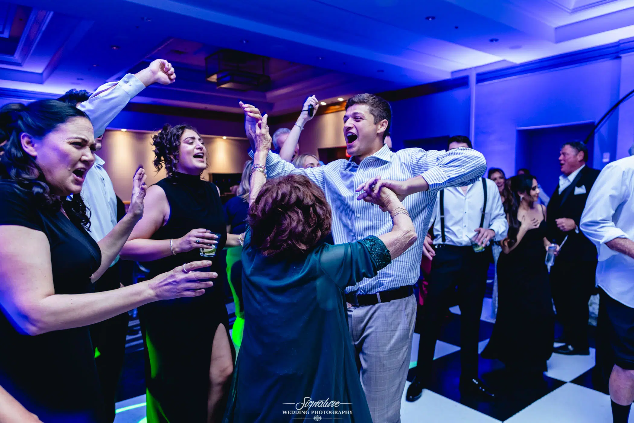 Image by Signature Wedding Photography A group of people enthusiastically dancing and celebrating at an indoor event, with colorful lighting and energetic expressions on their faces. Some have their arms raised and are laughing, creating a lively atmosphere.
