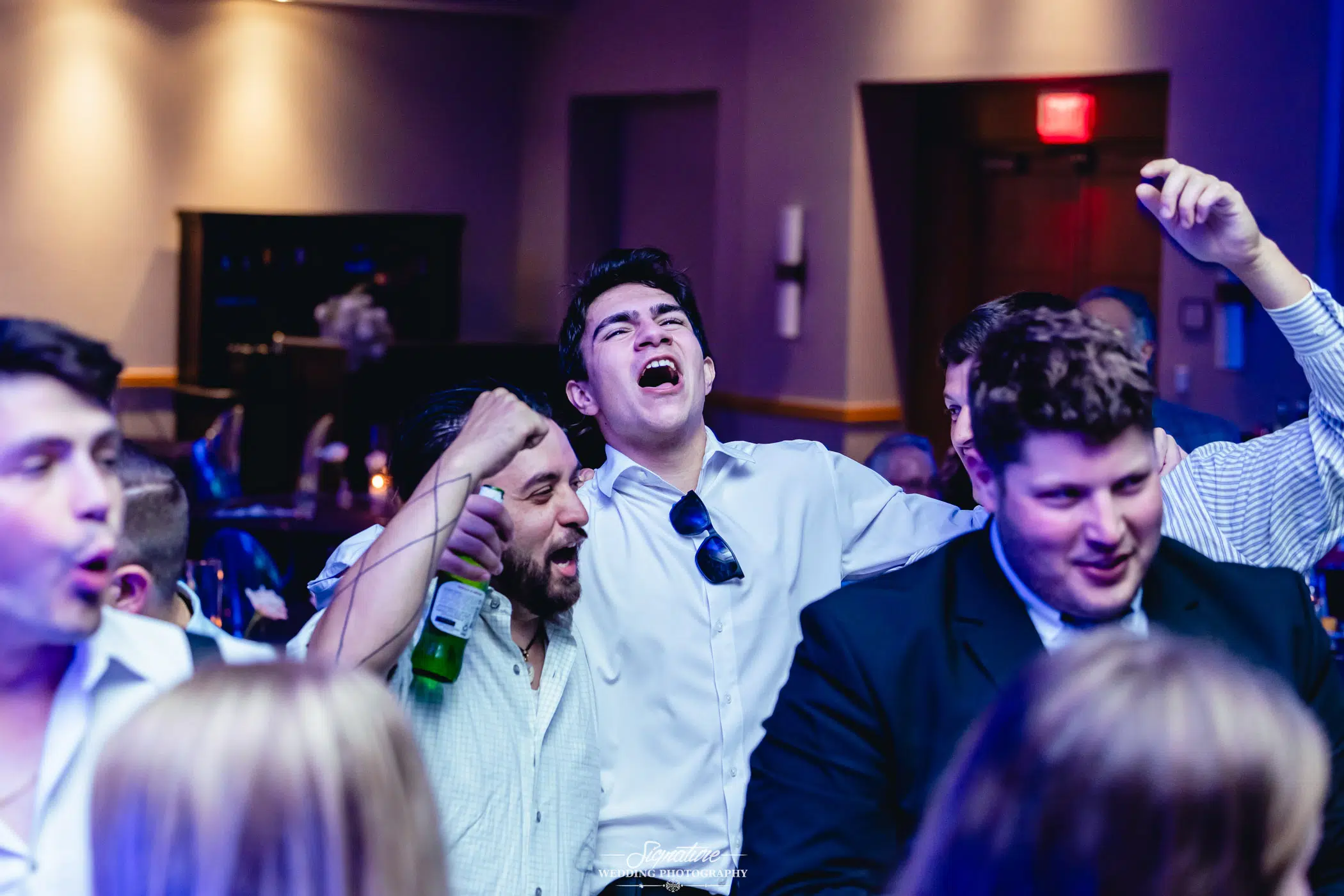 Image by Signature Wedding Photography A group of men in dress shirts and blazers energetically sing and cheer together at a party or event, with one man raising his arm and others holding drinks amid colorful lighting.