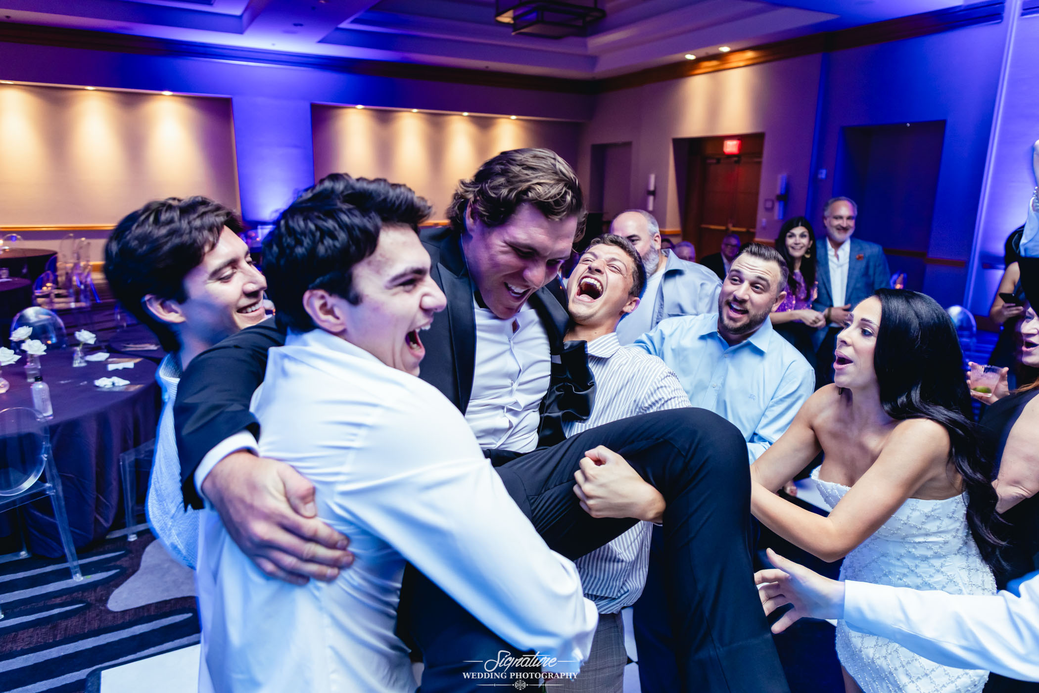 Image by Signature Wedding Photography A group of people at a celebration laughing and smiling as several men lift another man in the air. The scene is lively, with joyful expressions, a dimly lit room, and decorations in the background.