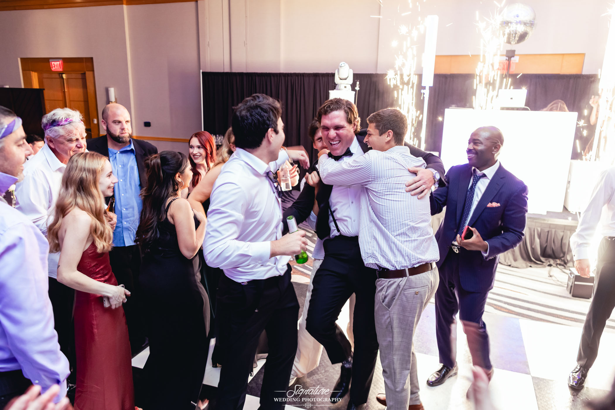 Image by Signature Wedding Photography A group of people in formal attire celebrate indoors, with two men hugging joyfully in the center. Others smile and watch, while sparklers light up in the background near a DJ booth. The mood is festive and lively.