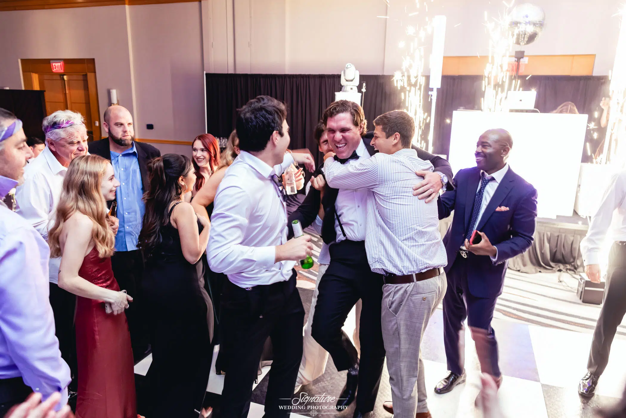 Image by Signature Wedding Photography A group of people in formal attire celebrate indoors, with two men hugging joyfully in the center. Others smile and watch, while sparklers light up in the background near a DJ booth. The mood is festive and lively.