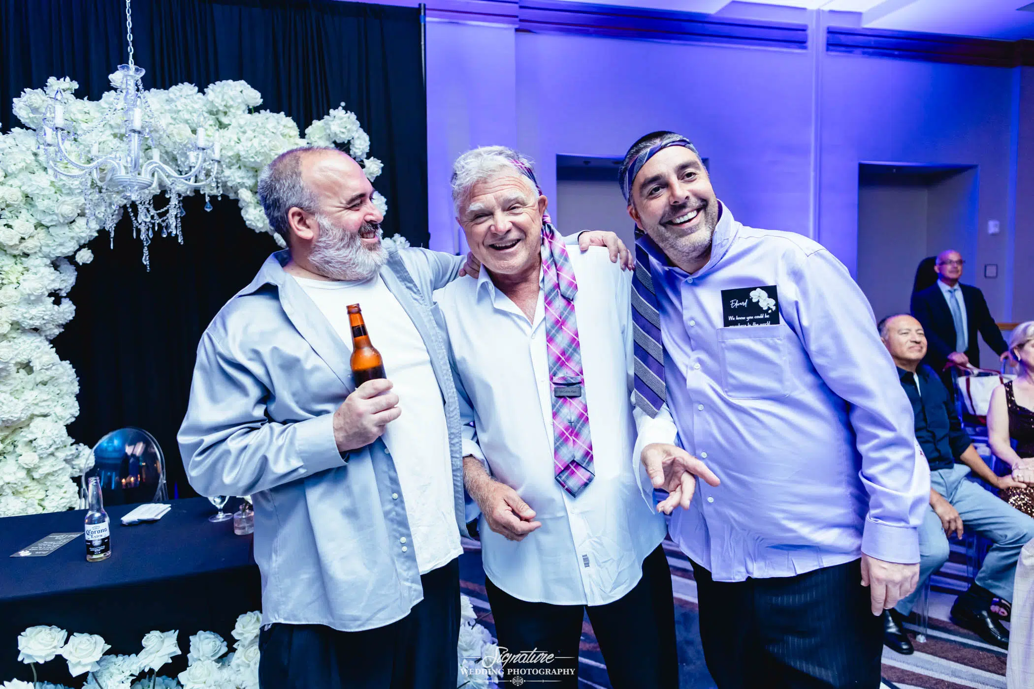 Image by Signature Wedding Photography Three older men smile and pose together at a celebration. One holds a beer, another wears a tie draped around his neck, and the third has a name tag and headband. White flowers and a chandelier decorate the background.