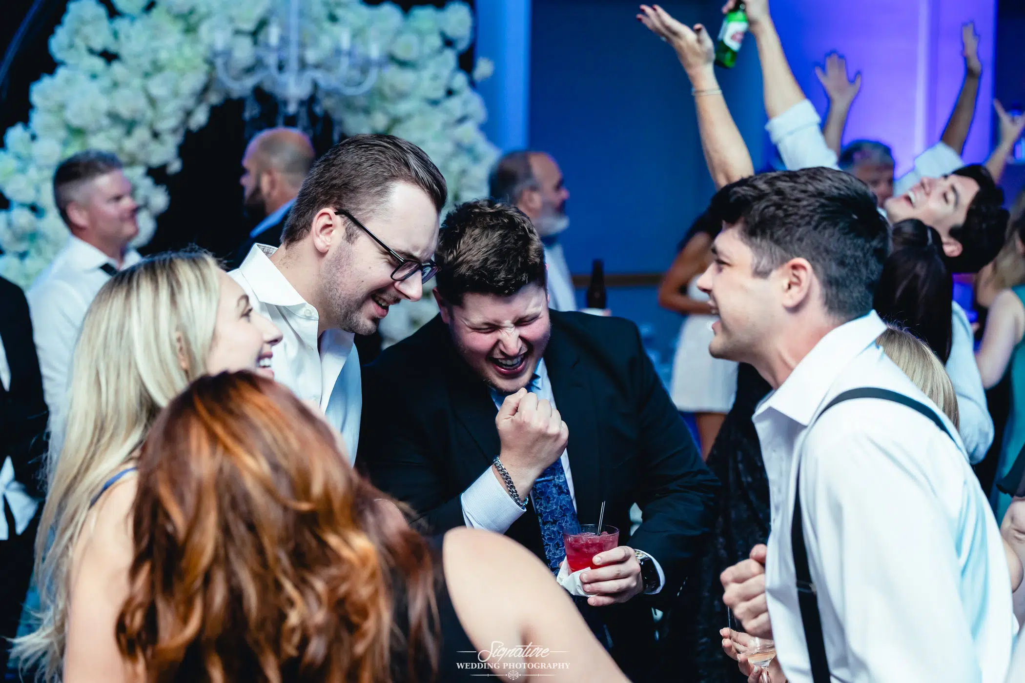 Image by Signature Wedding Photography A group of people in formal wear laugh and talk closely together on a lively dance floor at a celebration, with others dancing and cheering in the colorful background.